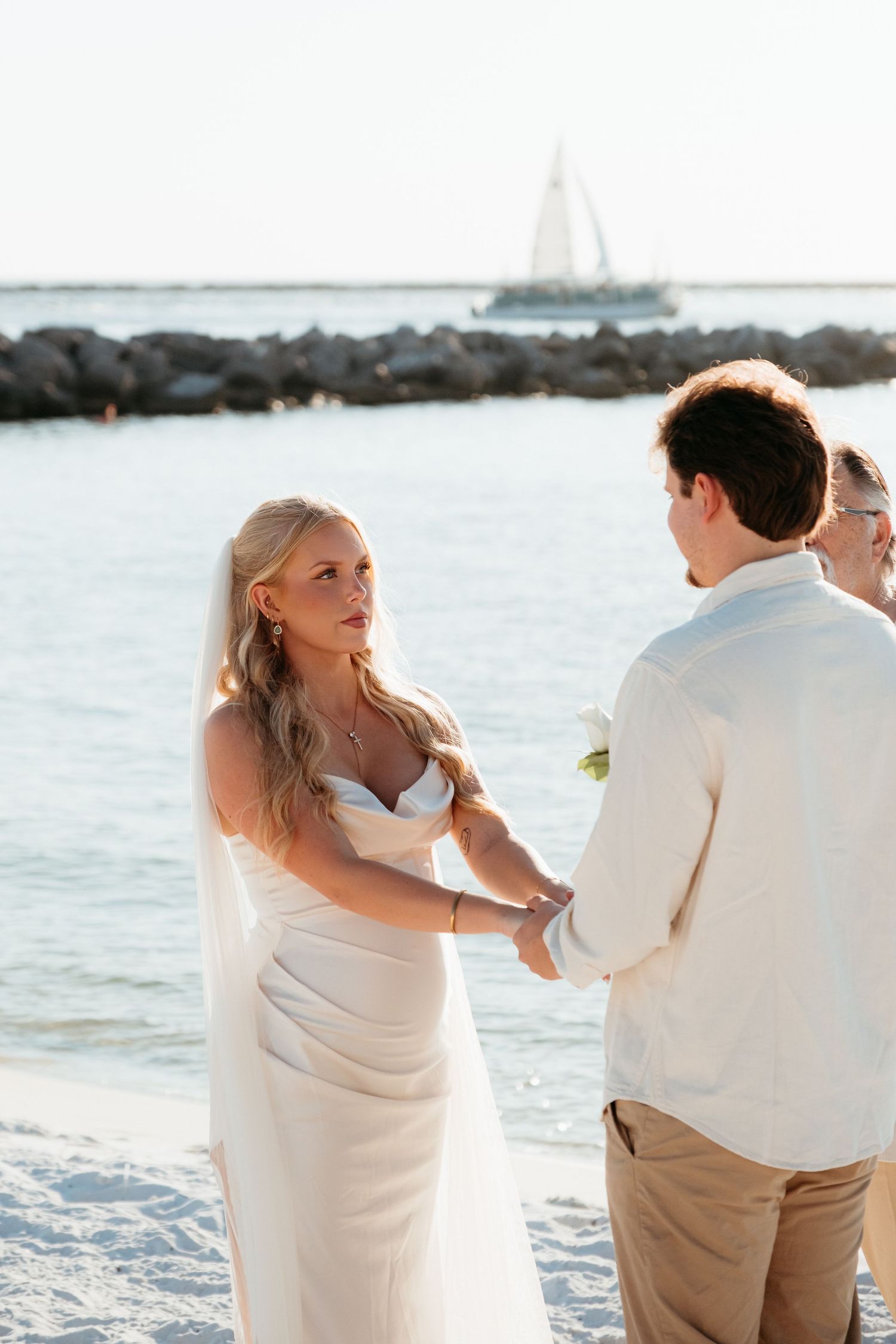 A couple in white wedding attire stands by ocean rocks with sailboat in background.