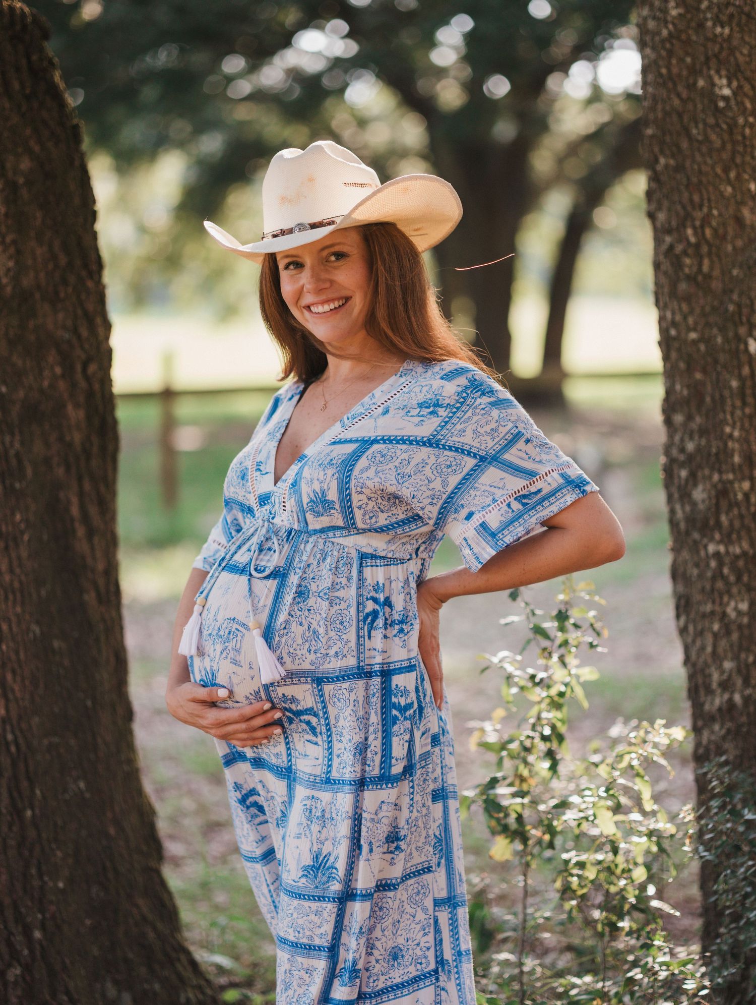 Woman in blue patterned dress and cowboy hat posing between trees.