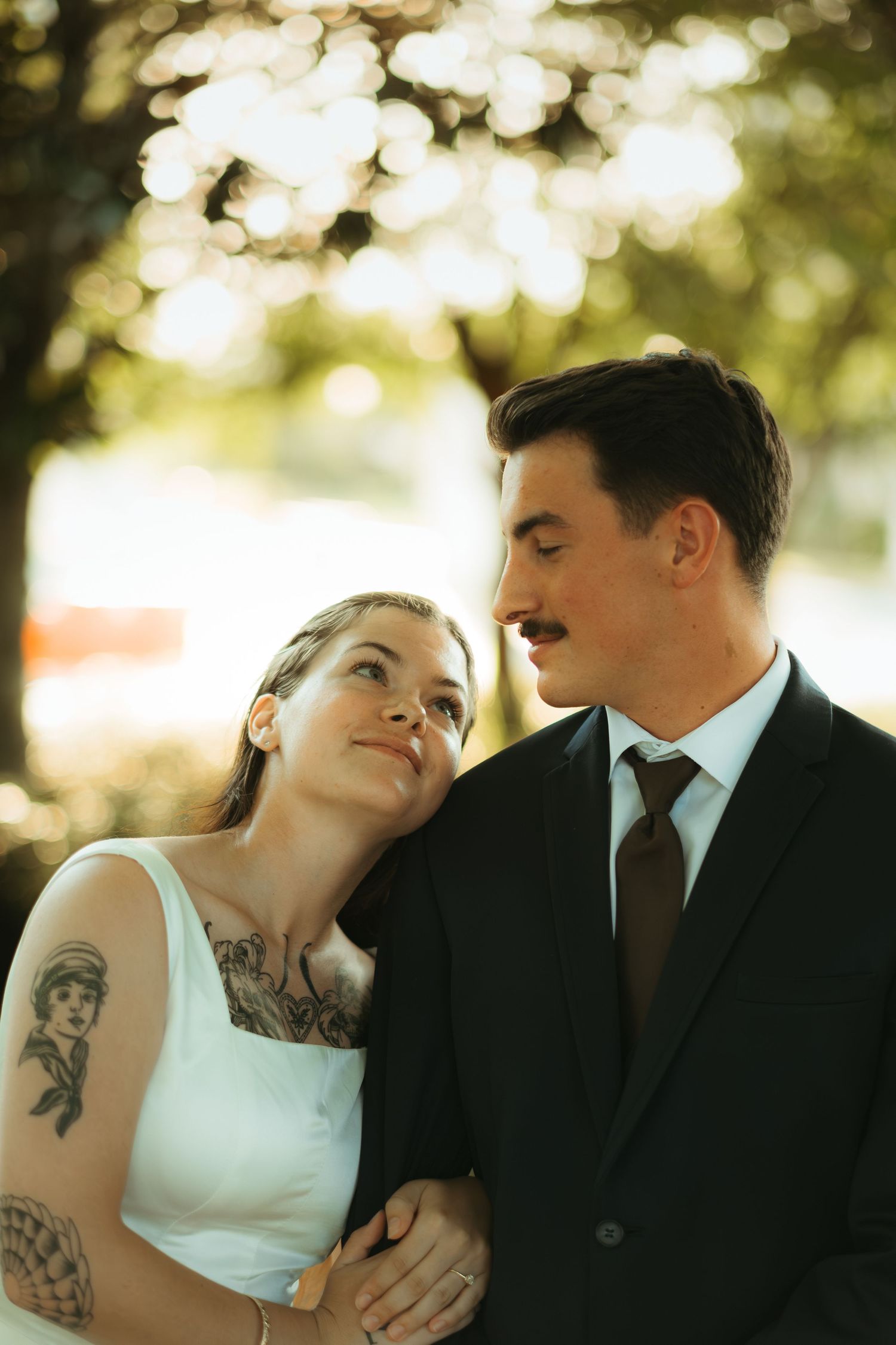 A couple in wedding attire poses together outdoors with soft bokeh lighting in trees.