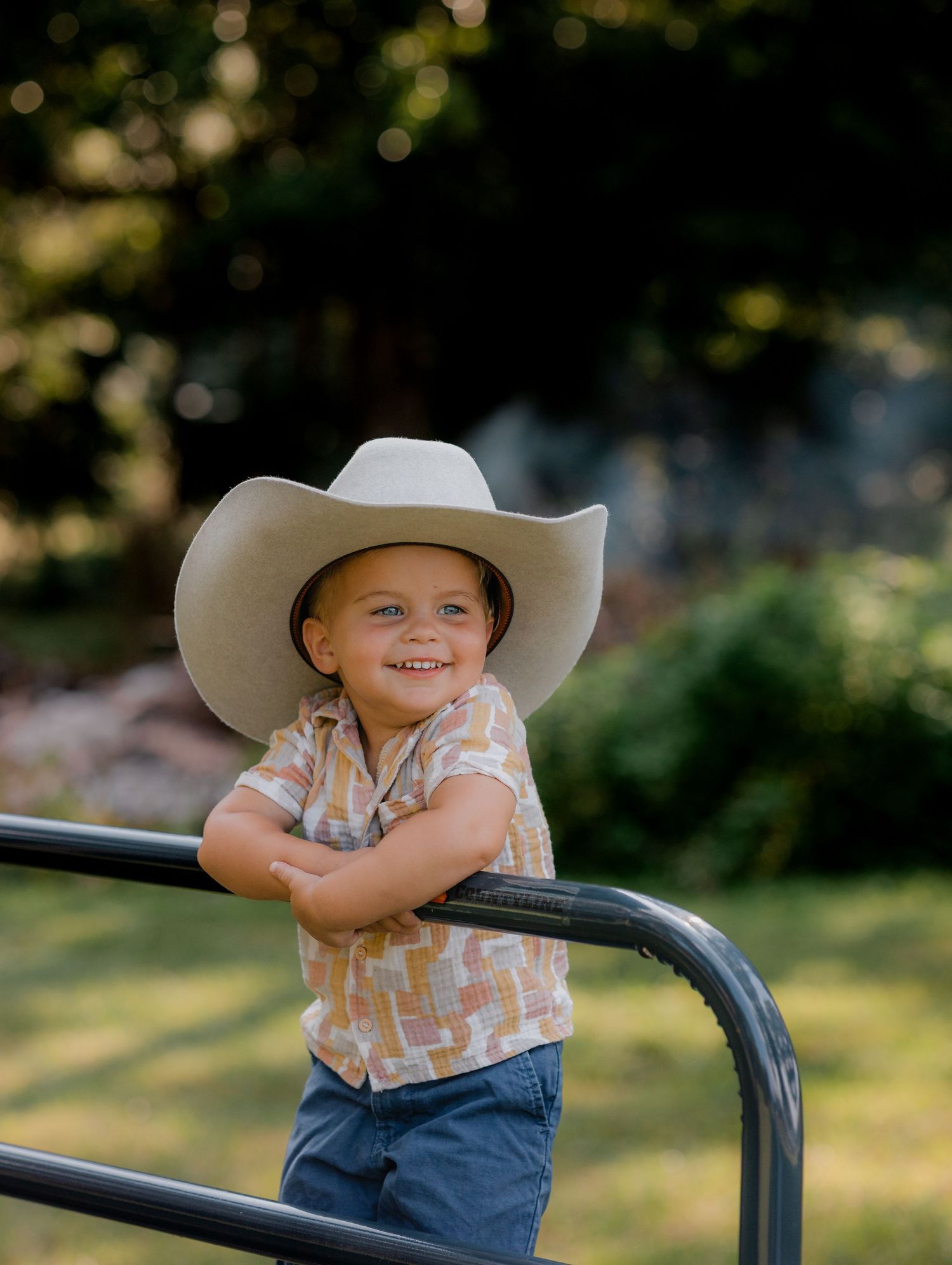 Child wearing western hat and casual clothes posing outdoors with natural bokeh background.