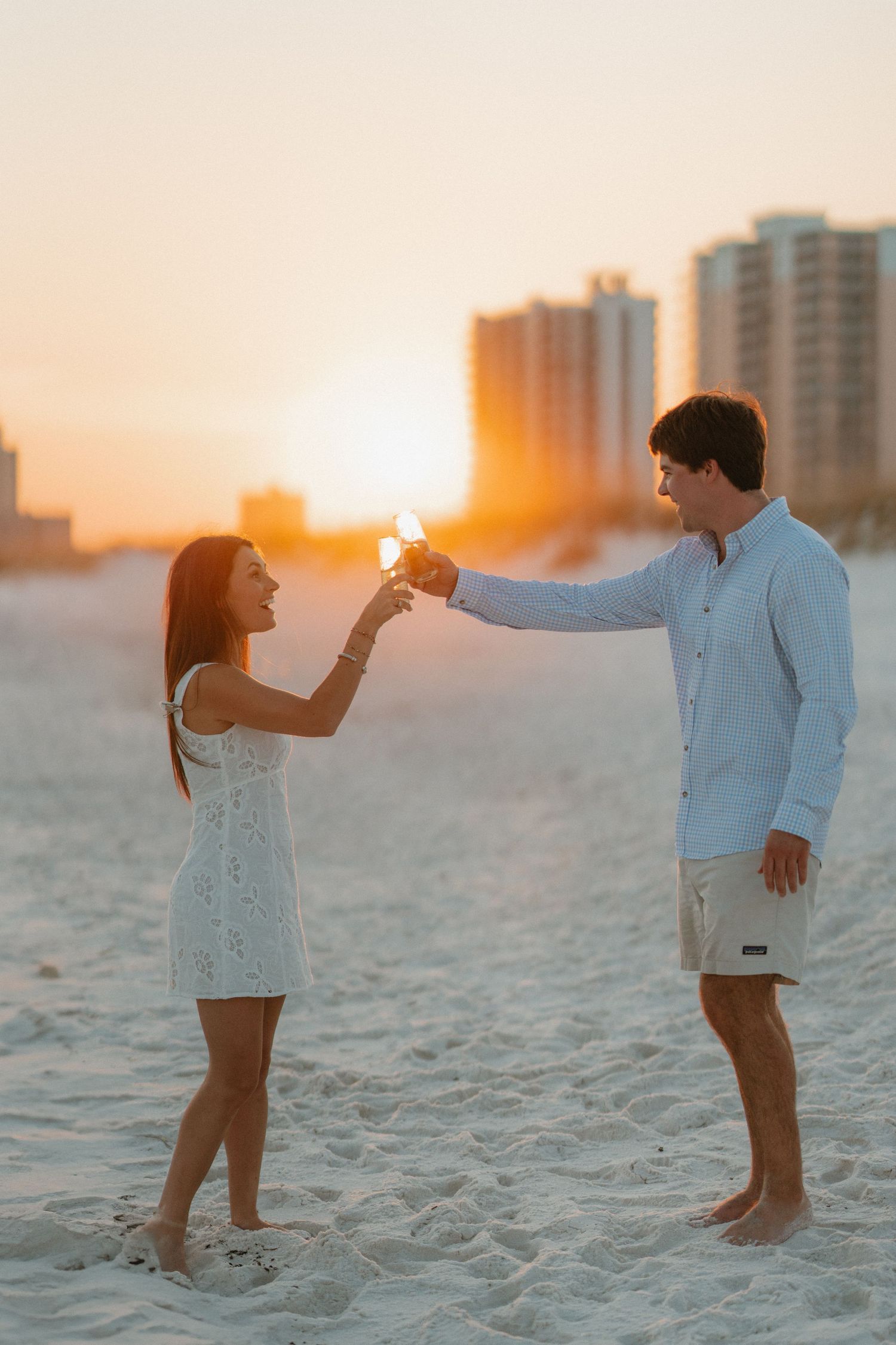 Couple playing and dancing on white sand beach at sunset with cityscape in background.