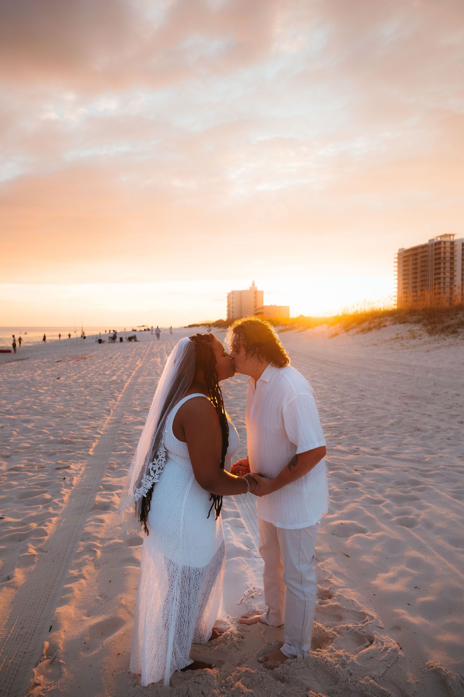 A couple shares an intimate moment on beach at sunset with high-rise buildings in background.