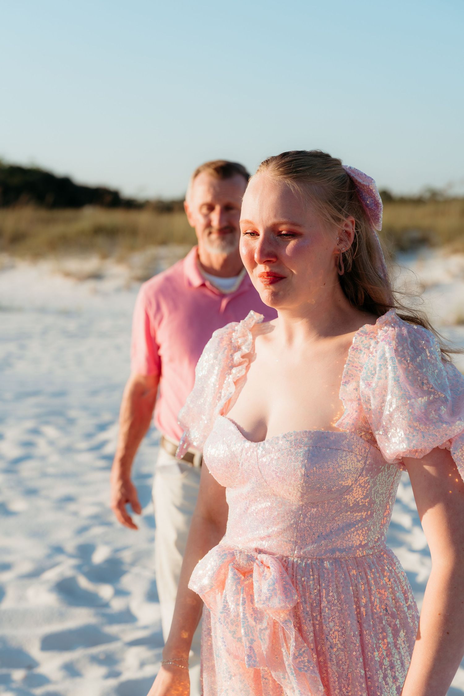 A couple in coordinating pink outfits poses together on a white sandy beach at sunset.