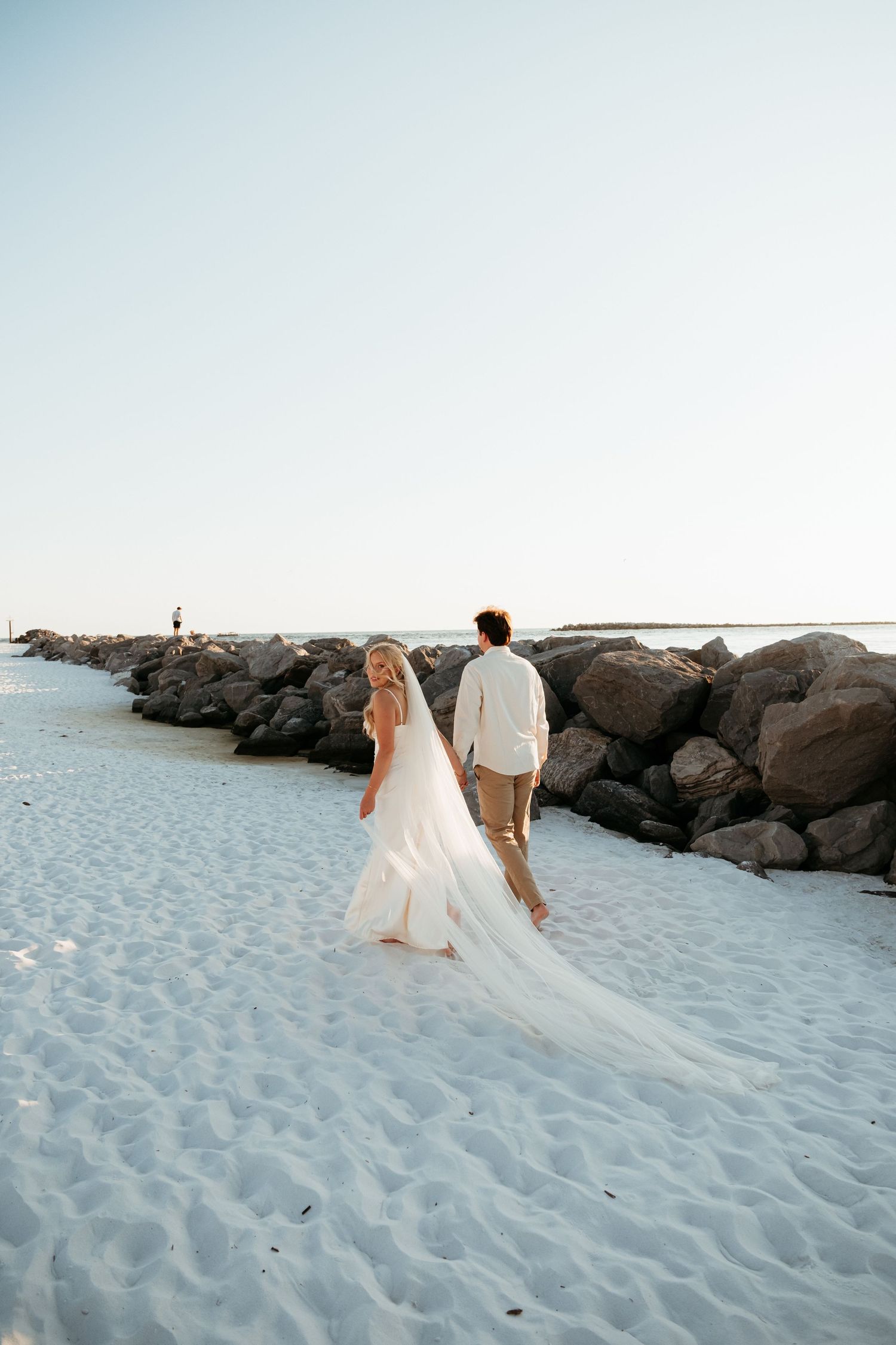 A couple walking along a rocky jetty on a white sandy beach.