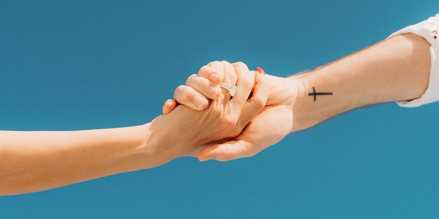Two hands clasped together against a blue background, with one arm showing a small cross tattoo.