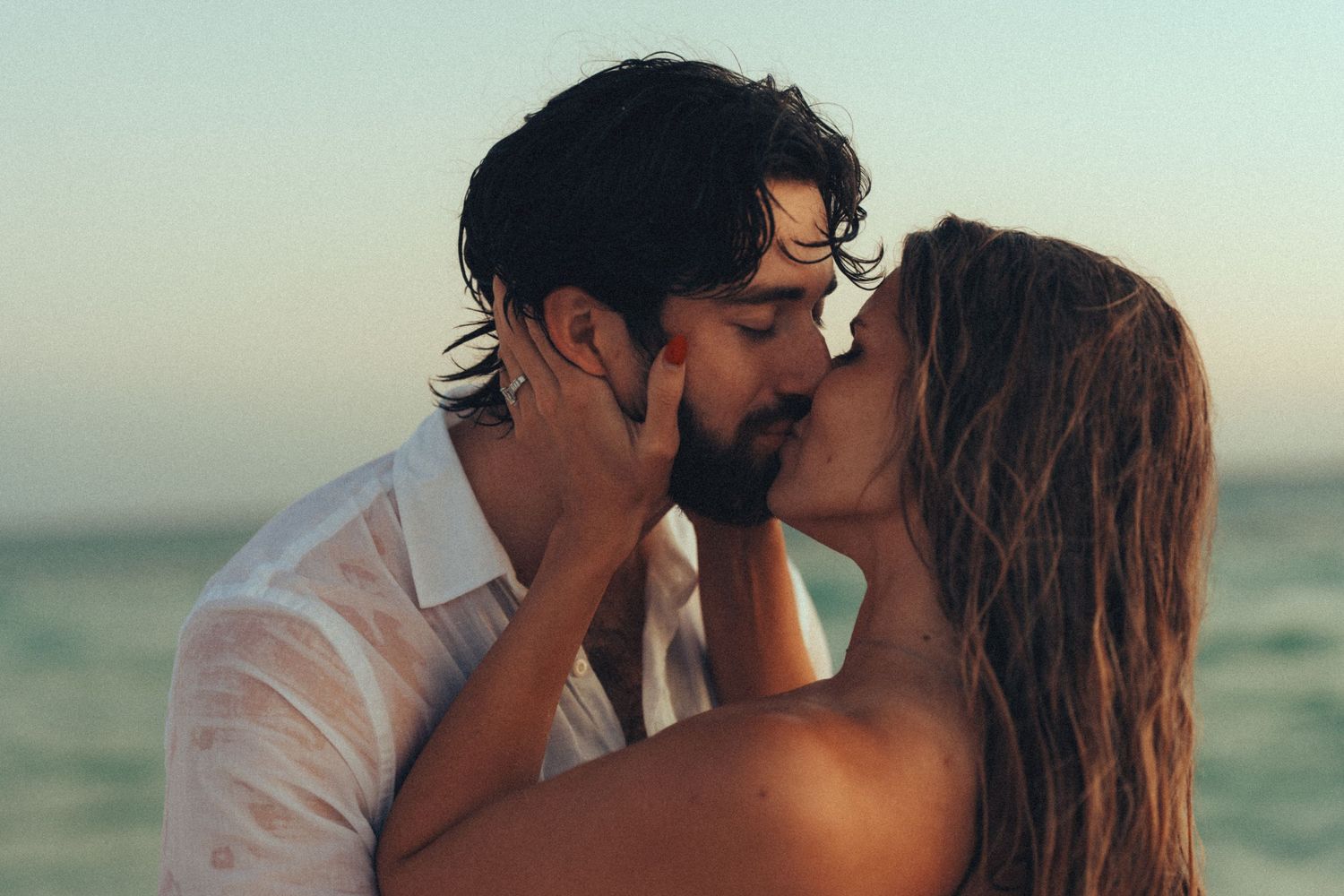 Romantic couple embraces and kisses on a beach at sunset with ocean waves in the background.