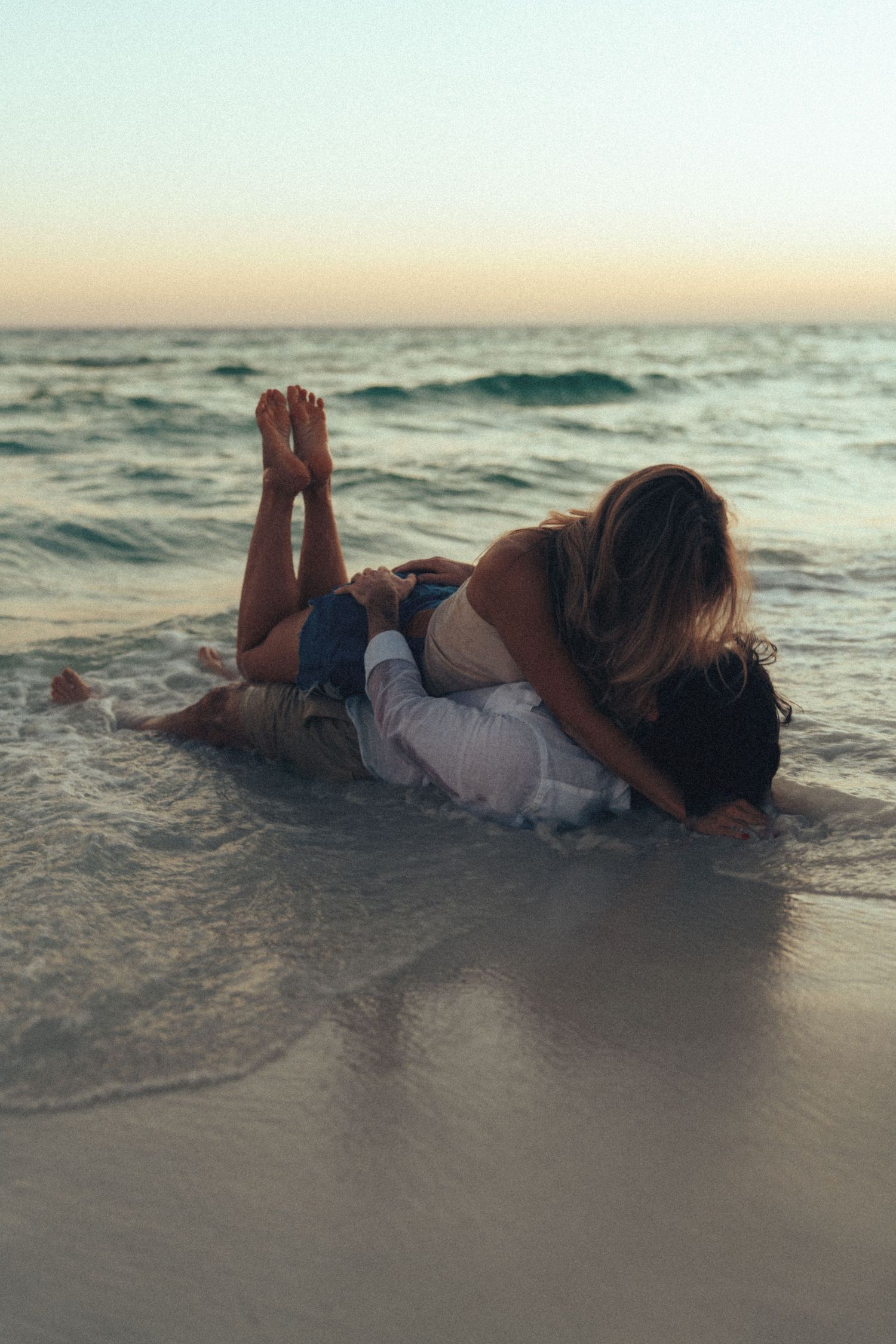 Person lying in shallow ocean waves at sunset wearing white clothing and creating artistic beach photography.
