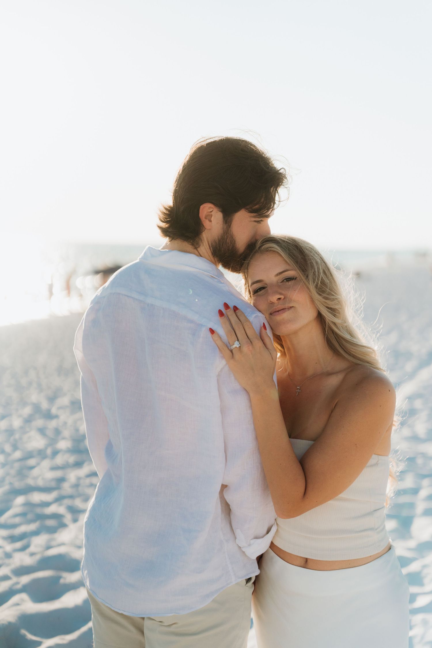 A couple embraces romantically on a bright sandy beach in white attire.