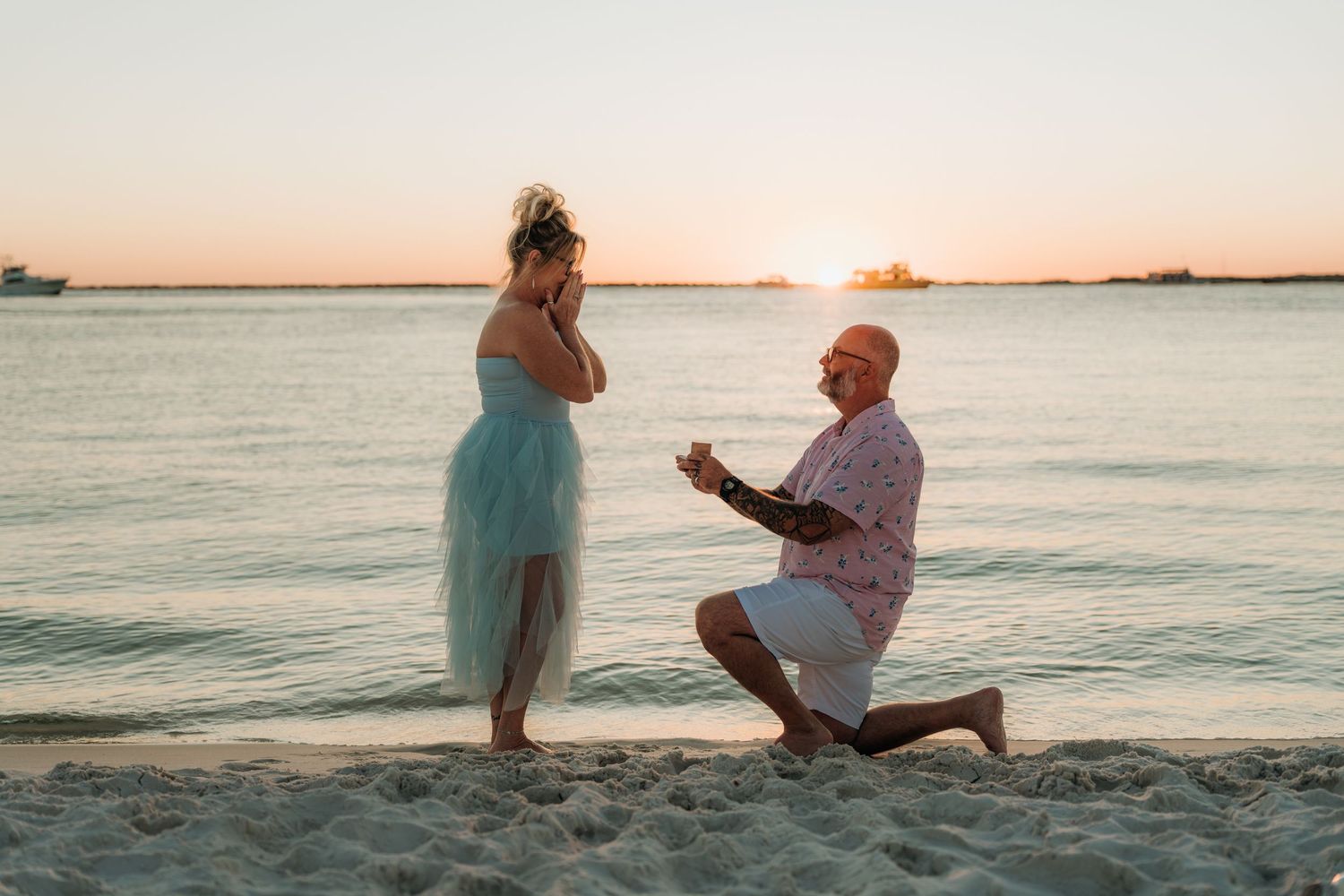 A romantic beach proposal at sunset with a person kneeling in front of their partner on the sandy shore.
