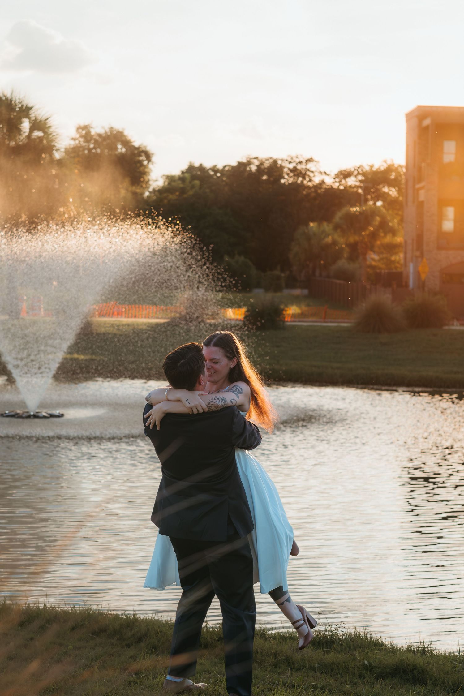 A couple embraces by a fountain at sunset with golden light streaming through trees.