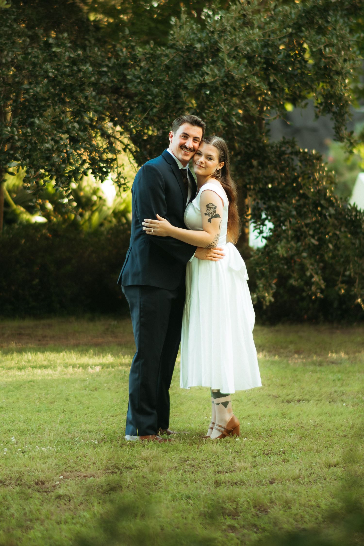 A couple in formal attire embraces on grassy lawn during an outdoor wedding photoshoot.