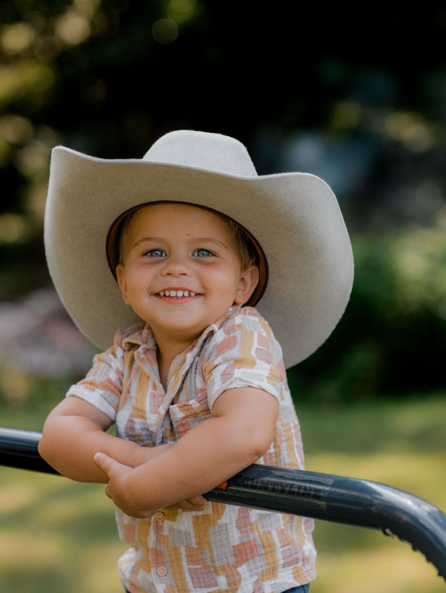 Young child in cowboy hat smiling while leaning on a railing in summer attire.