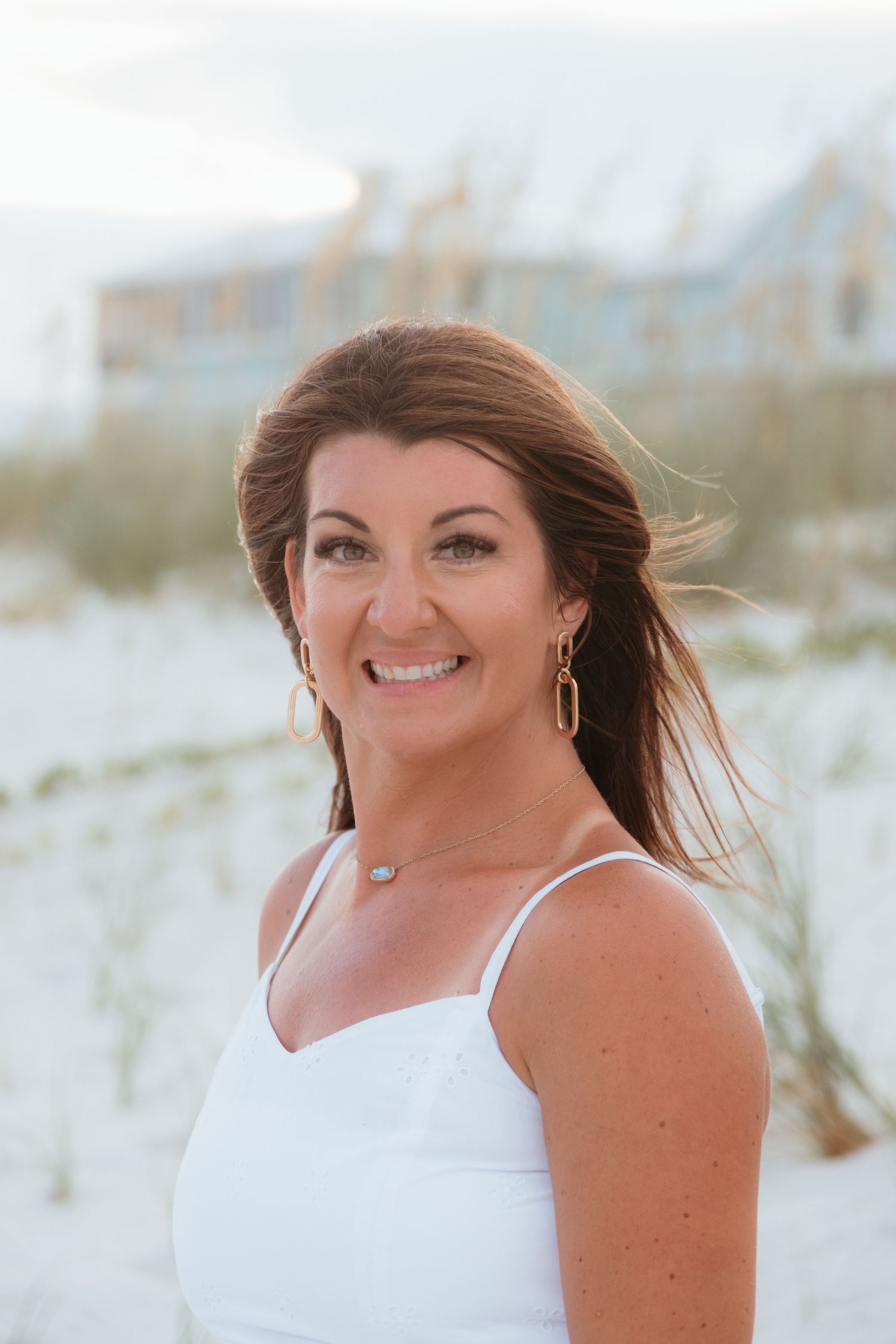 Woman in white sundress smiling on beach with sand dunes in background.