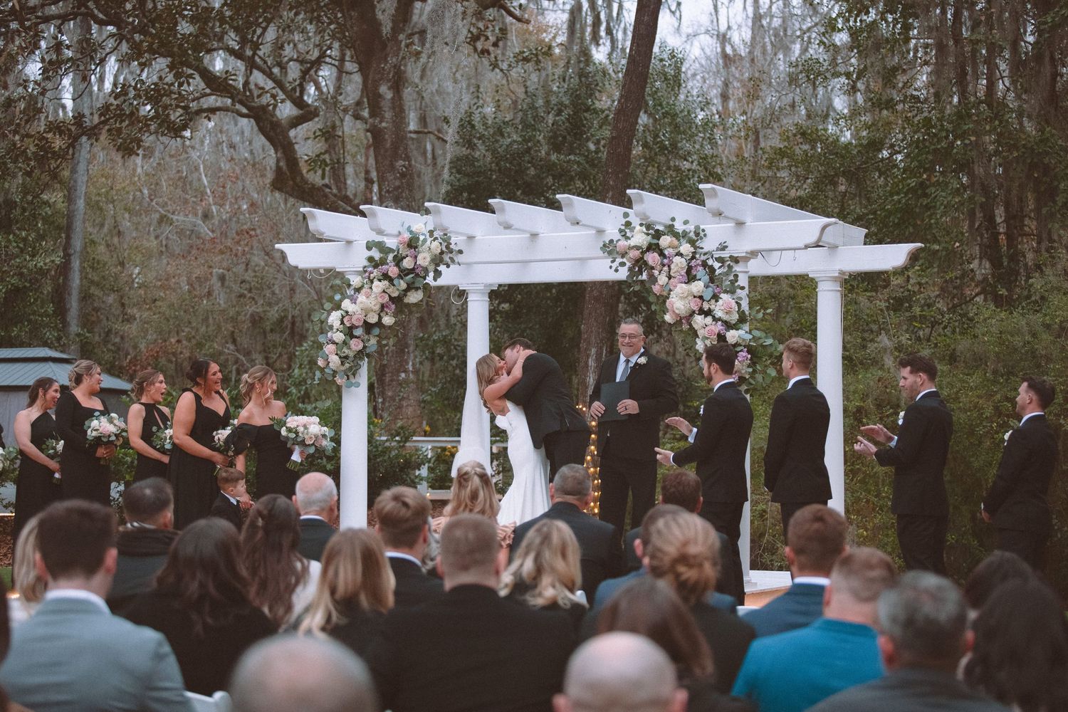 Bride and groom share first kiss under a floral-adorned white pergola at an outdoor wedding ceremony.