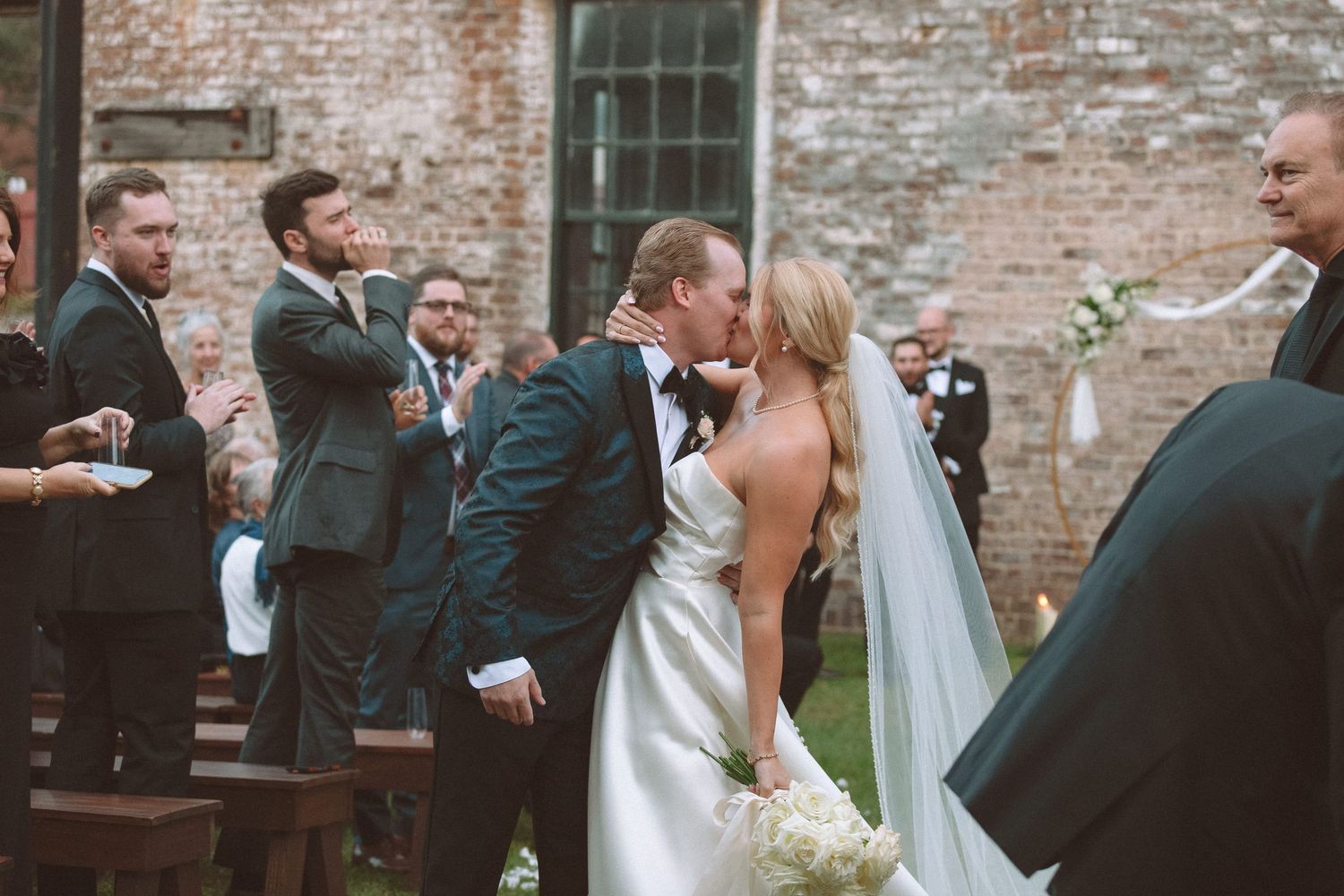 Bride and groom sharing first kiss at outdoor wedding ceremony surrounded by brick walls and cheering guests.