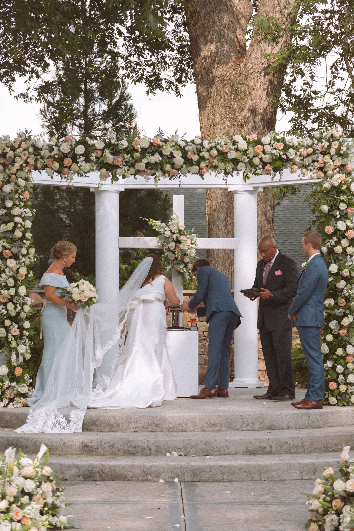 Bride and groom exchange vows under white floral arch gazebo at outdoor wedding ceremony.