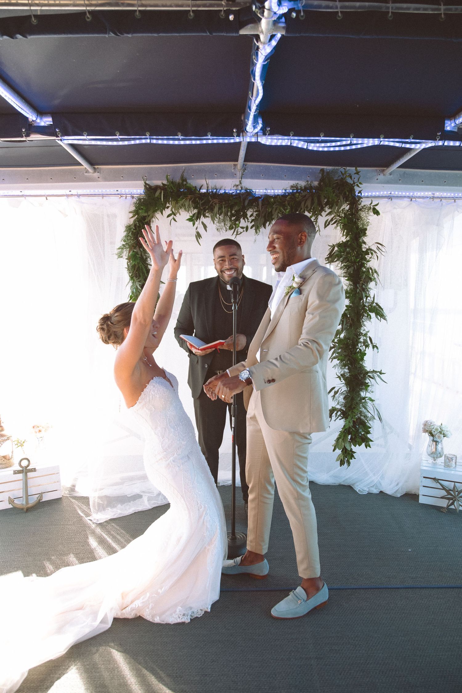 Bride raises hands in joy as groom smiles during wedding vows under lush greenery arch.