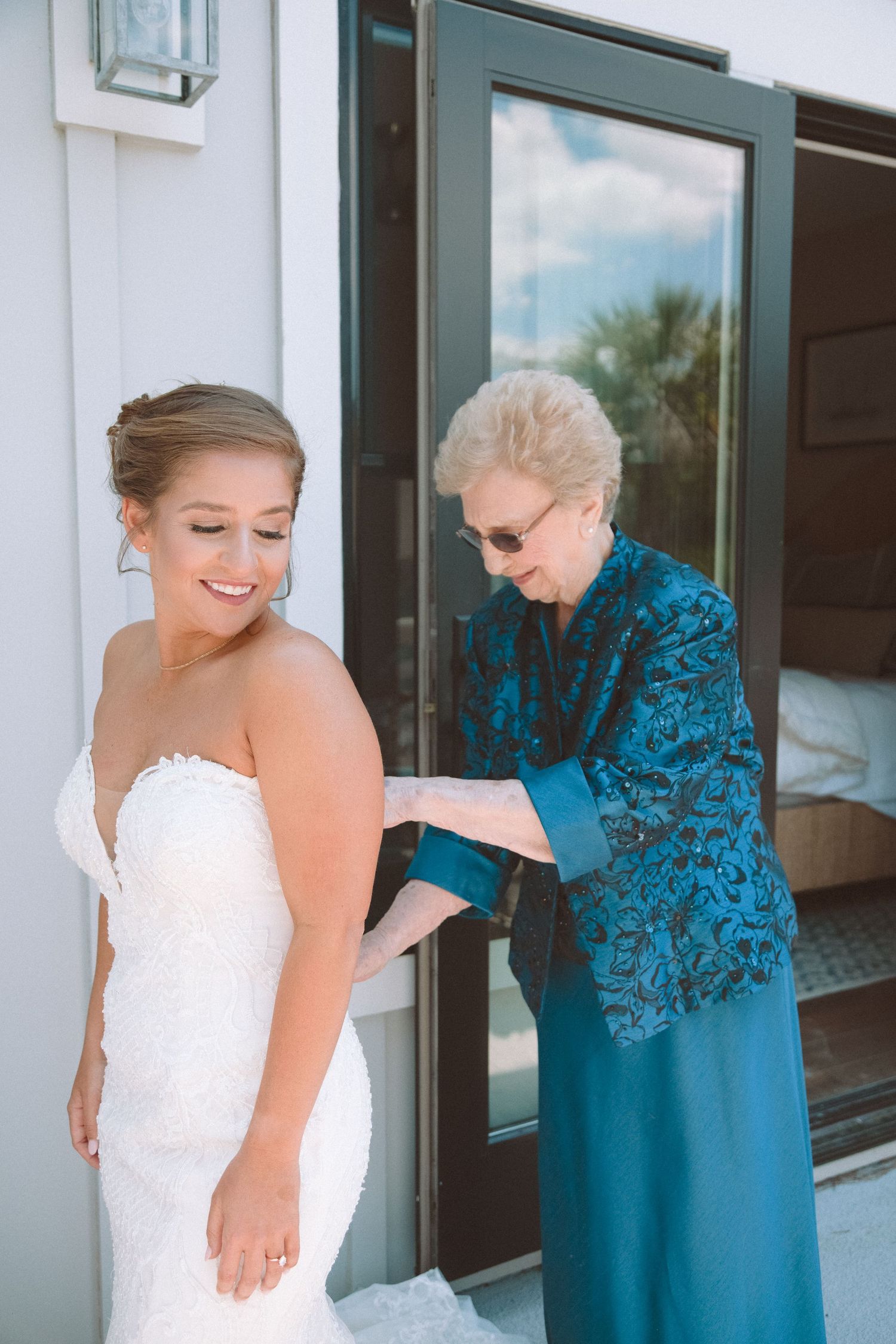 Grandmother fastening the back of a brides fitted white lace wedding dress outdoors.