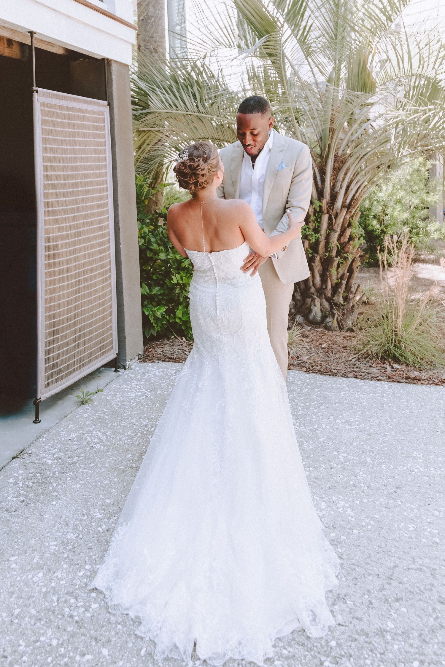 Groom admires bride's elegant lace mermaid wedding gown from behind during outdoor first look near palms.