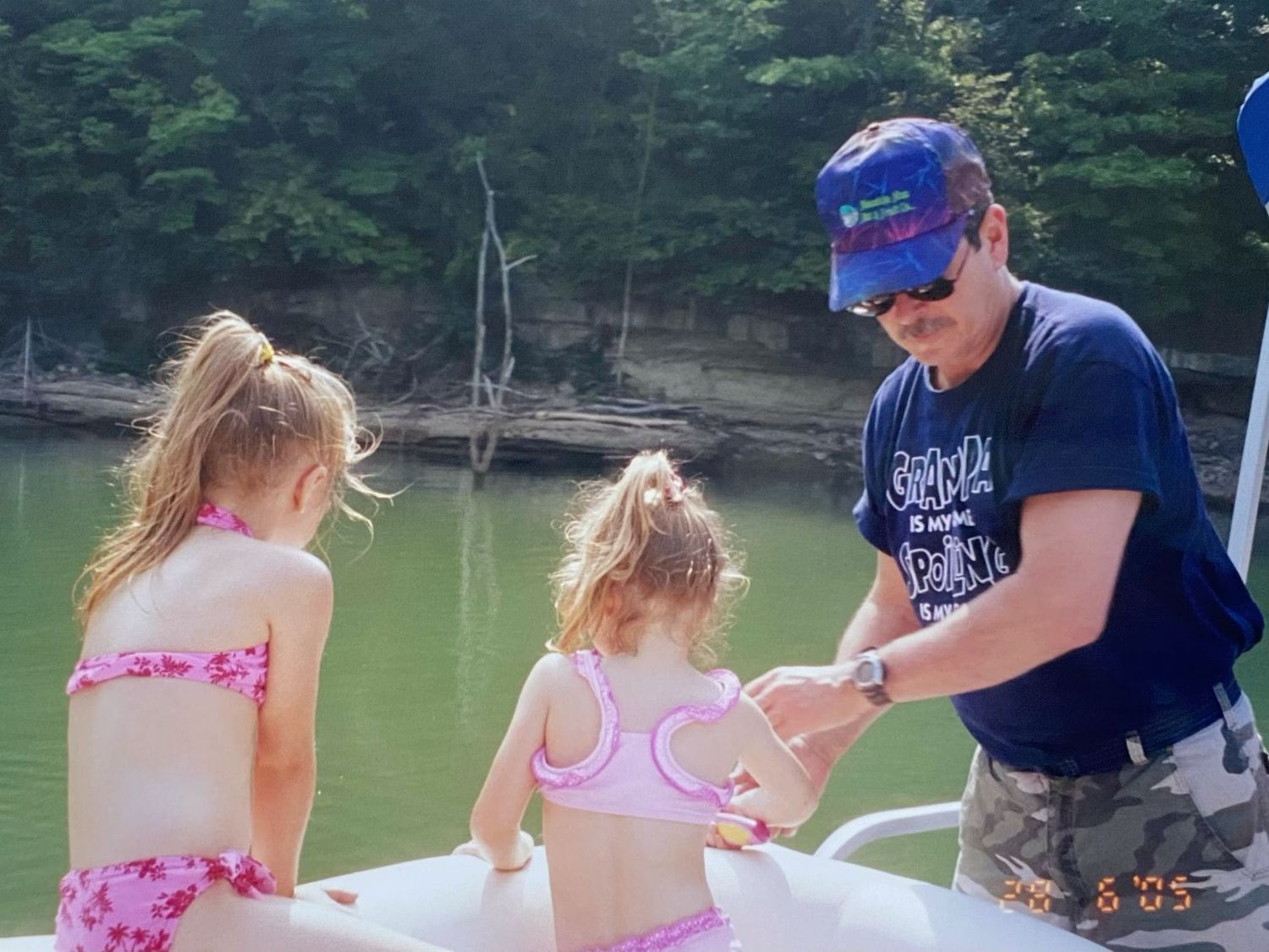 A man in a blue cap helping two young girls in pink swimsuits on a boat near a wooded lake.