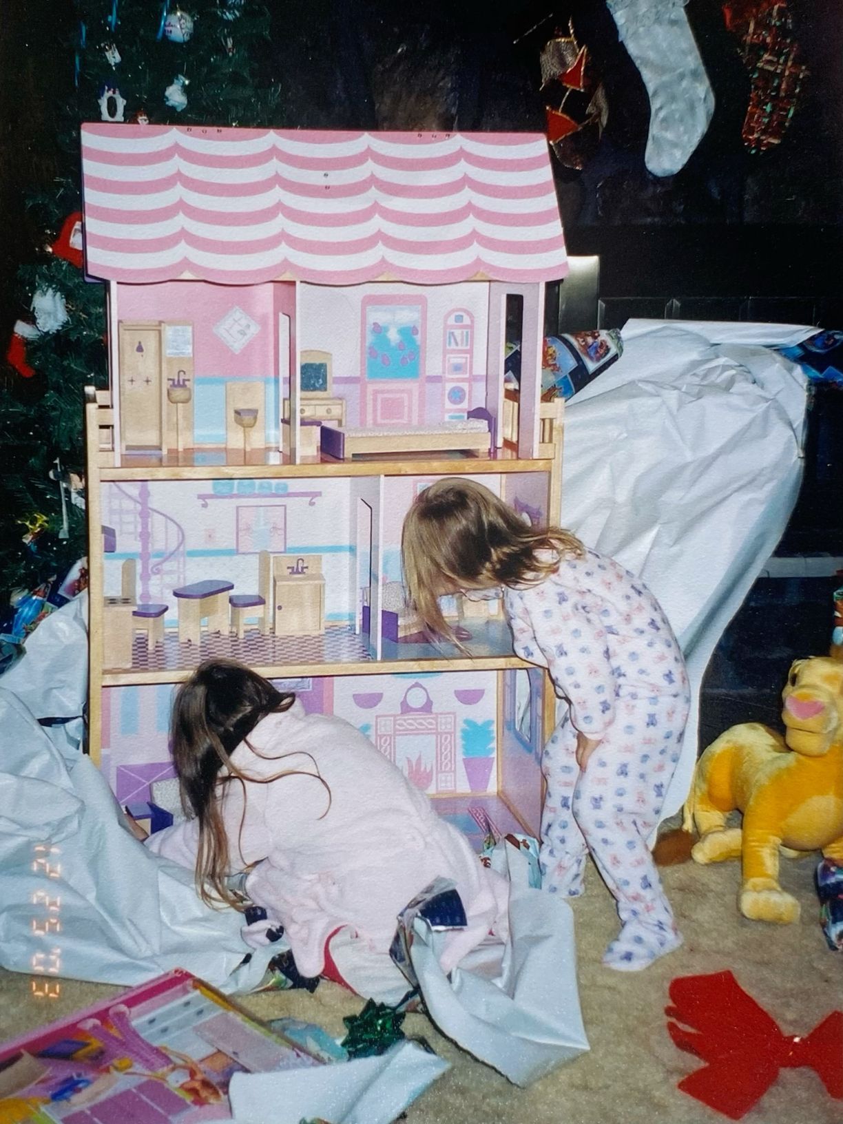 Two young girls in pajamas excitedly exploring a large pink wooden dollhouse on Christmas morning.