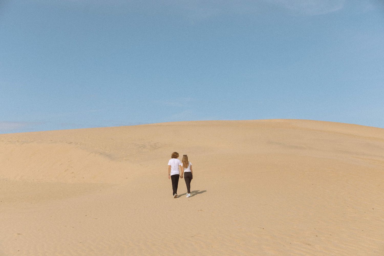Two people holding hands walking up a vast sandy desert dune under clear blue sky.