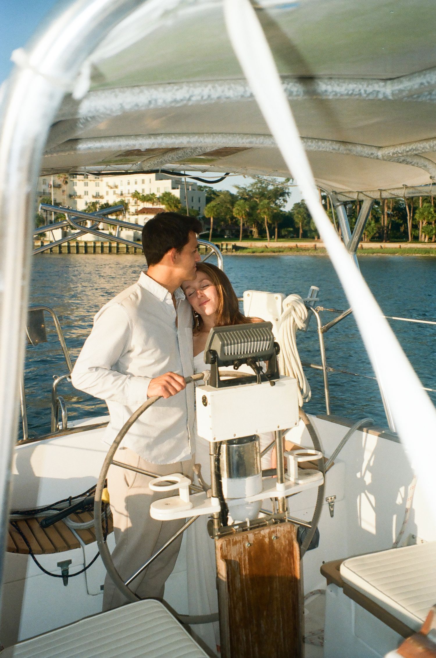 Romantic couple embracing at the helm of a white sailboat on calm blue waters near a tropical shoreline.
