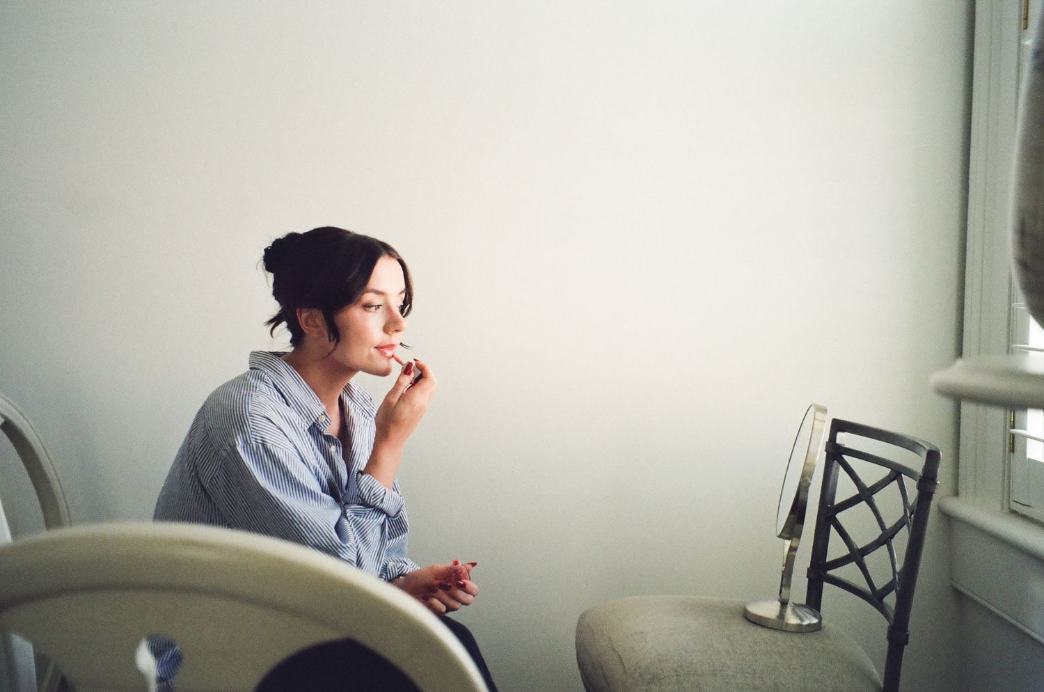 A thoughtful young woman sits by a window in a blue shirt, resting her chin on her hand in a minimalist room.