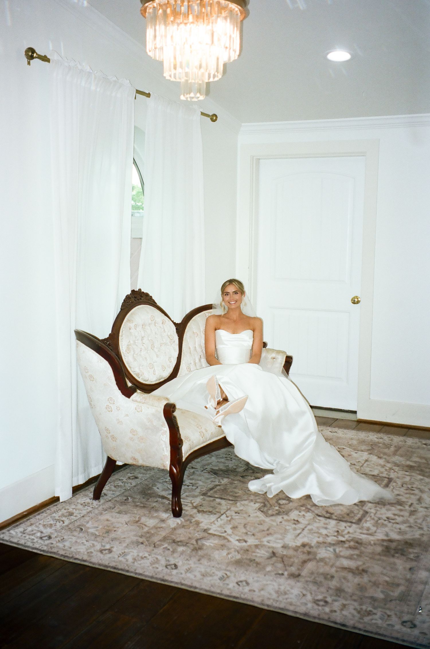 Radiant bride in strapless white gown seated on ornate vintage settee beneath crystal chandelier.