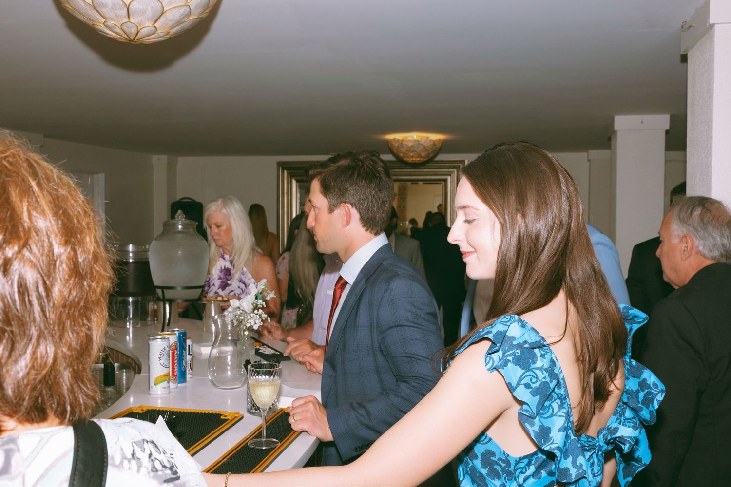 Wedding guests mingle at an elegant indoor bar reception, with a young woman in a blue floral dress.