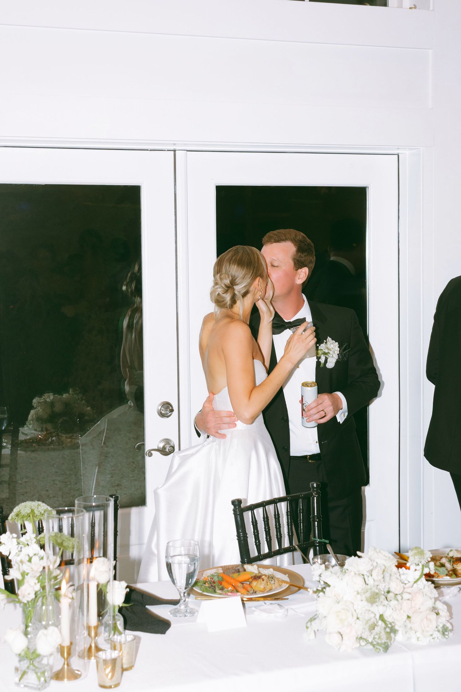 Bride and groom share a kiss at wedding reception table adorned with white floral centerpieces and candles.