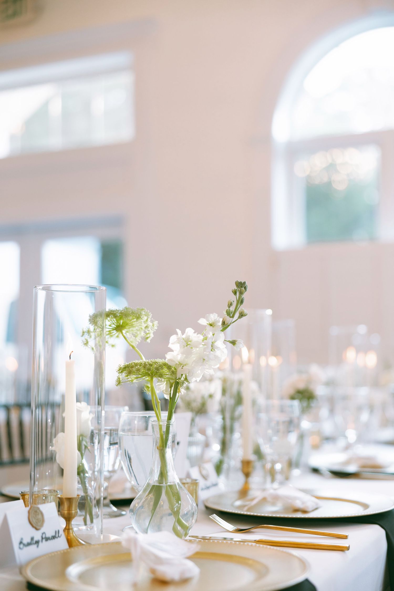Sophisticated wedding reception table with white and green floral arrangements, candles, and gold accents in bright venue.