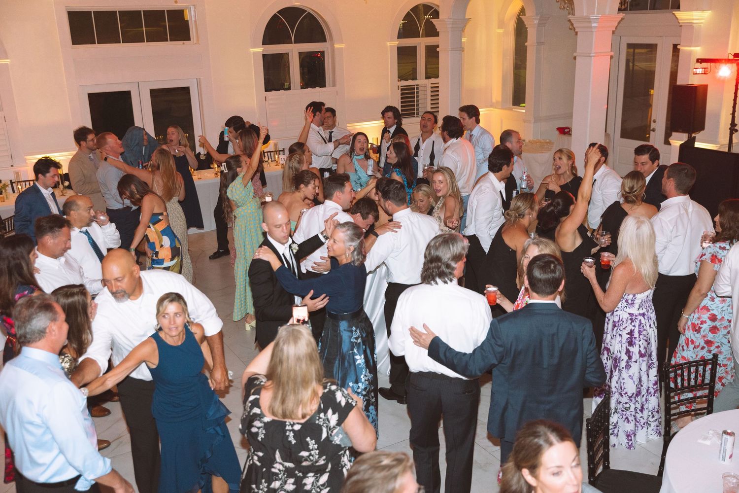 Crowded wedding reception dance floor with guests dancing in elegant attire inside a bright white venue.