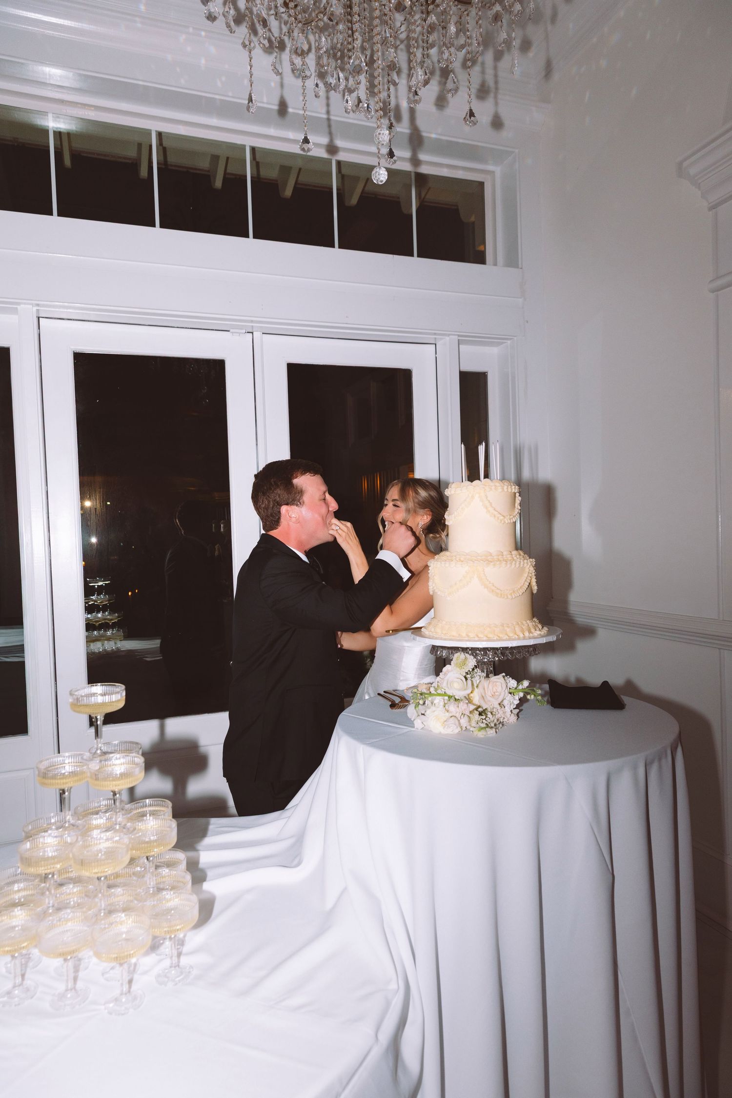 Bride feeding groom wedding cake beside champagne tower at elegant white wedding reception.