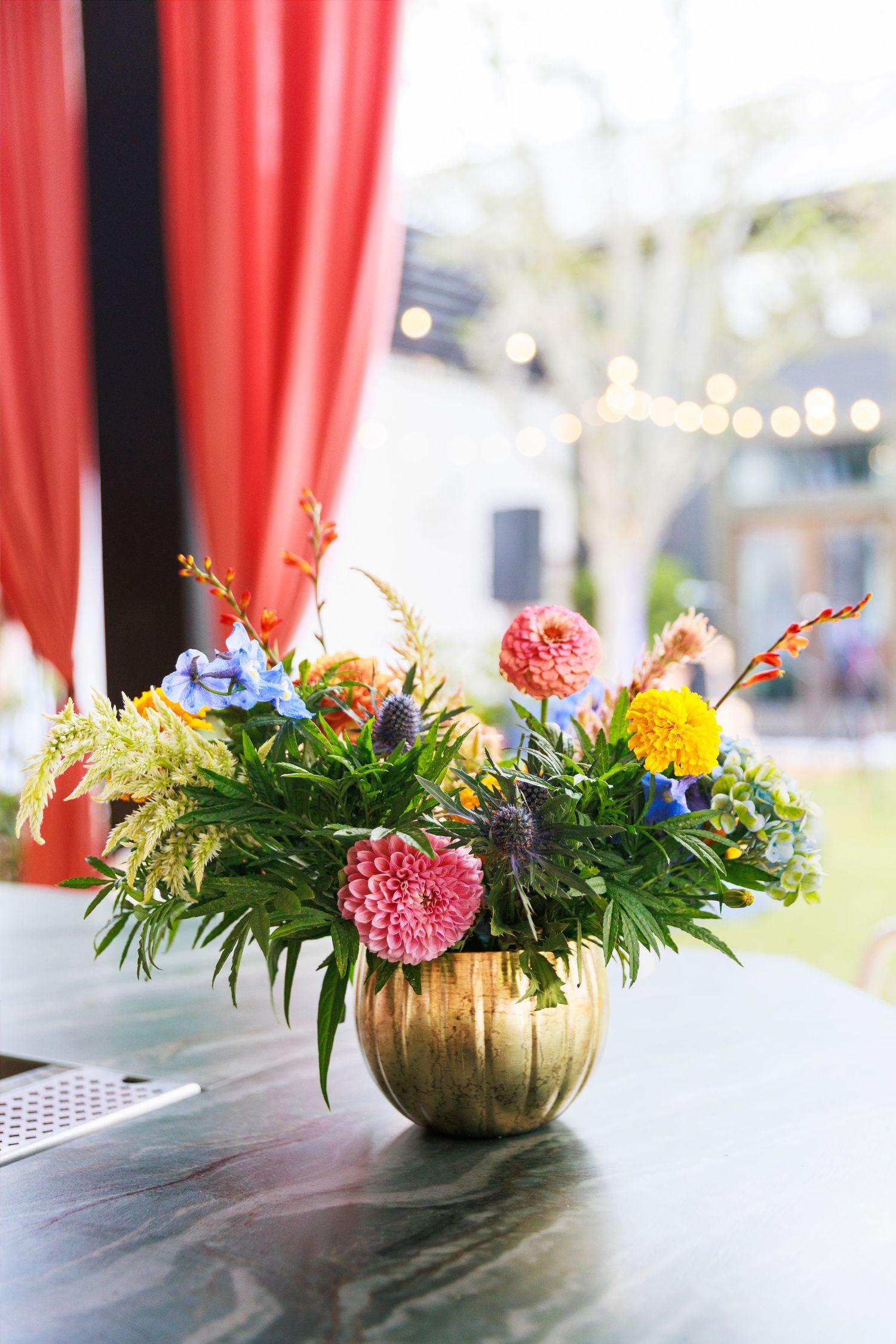 Vibrant mixed floral arrangement in a gold vase with dahlias and wildflowers on a marble table.