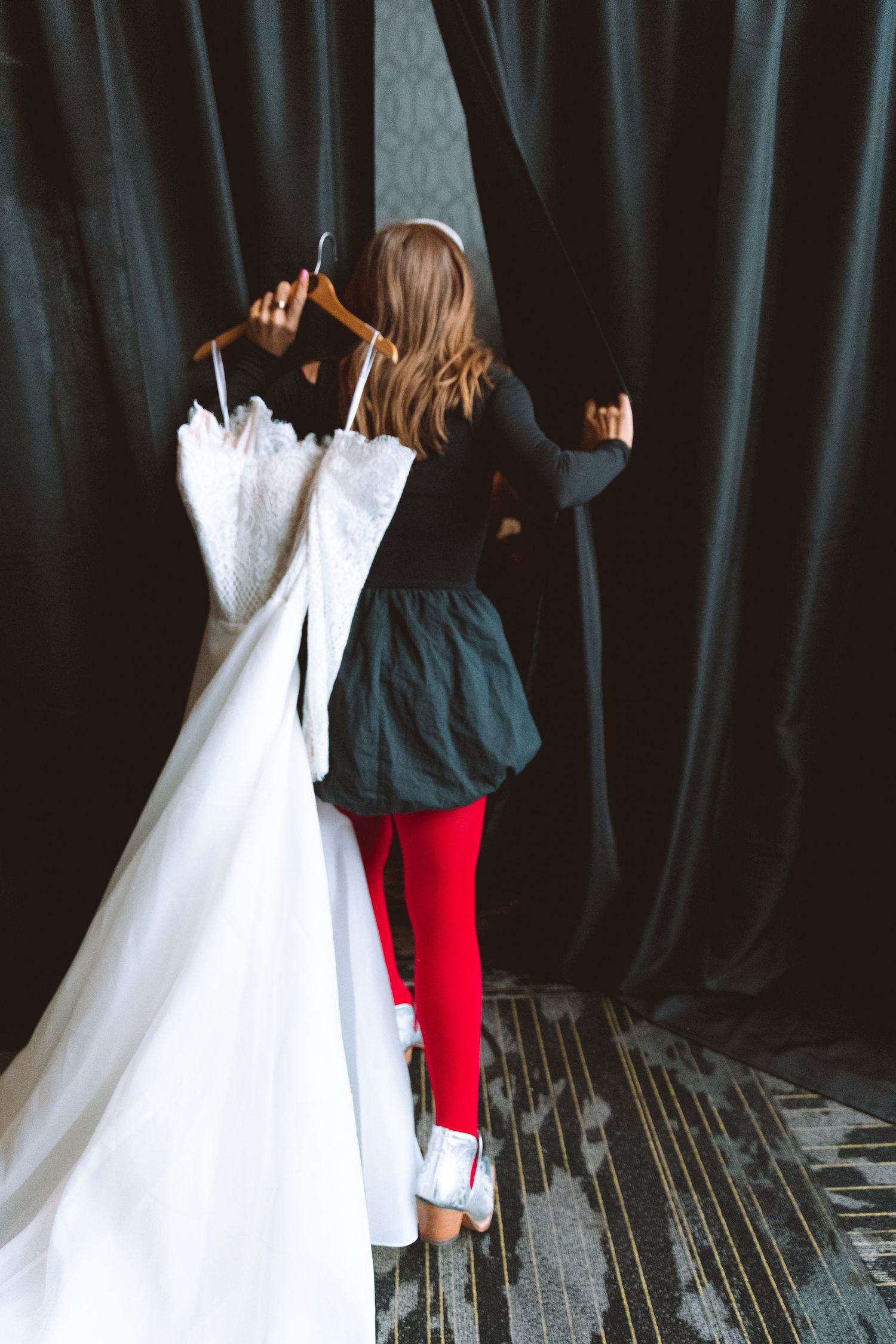 Woman holds white wedding dress on hanger near black curtain backstage at bridal fashion event.