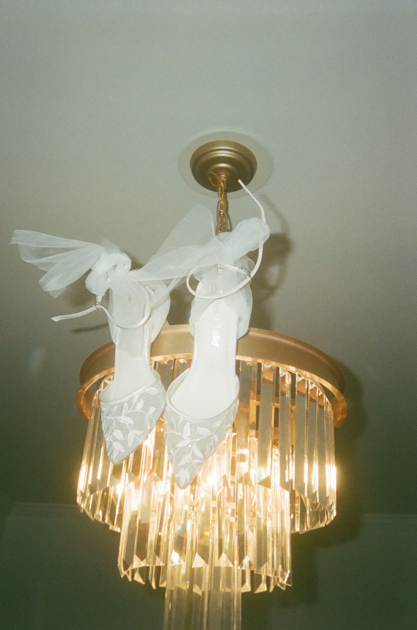 White bridal heels with floral details hanging from a gold crystal chandelier, elegant wedding shoe display.
