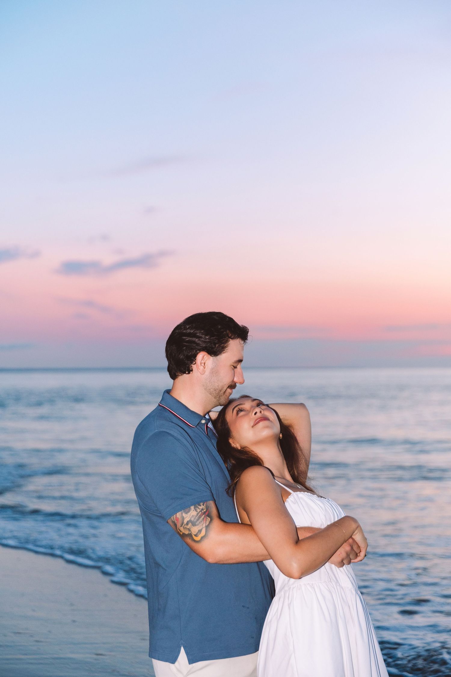 Couple embracing romantically on a beach at sunset with pink and blue sky over the ocean.