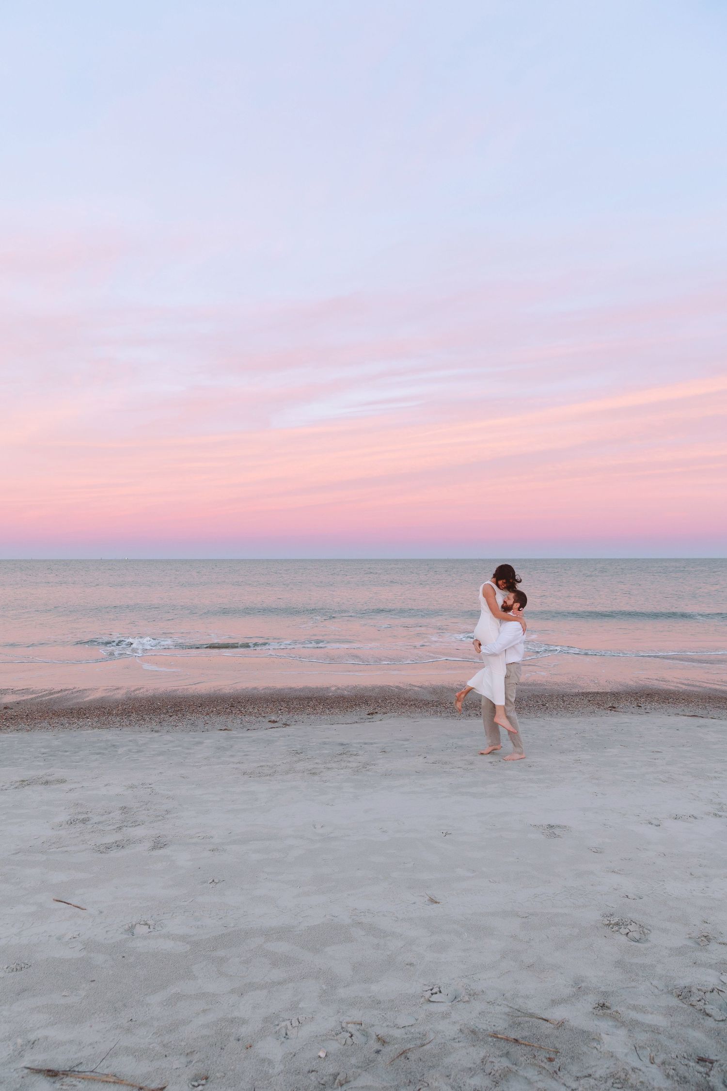 Couple embracing on a sandy beach at sunset with pink and blue sky reflecting over calm ocean waters.