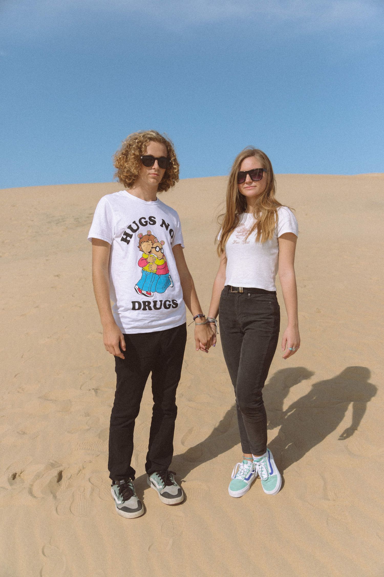 Young couple holding hands wearing white t-shirts standing on sandy desert dunes under blue sky.