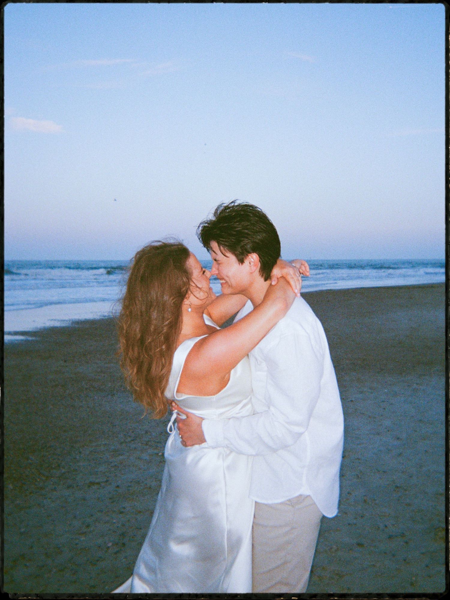 Romantic couple embracing and almost kissing on a beach at dusk, both wearing white outfits.