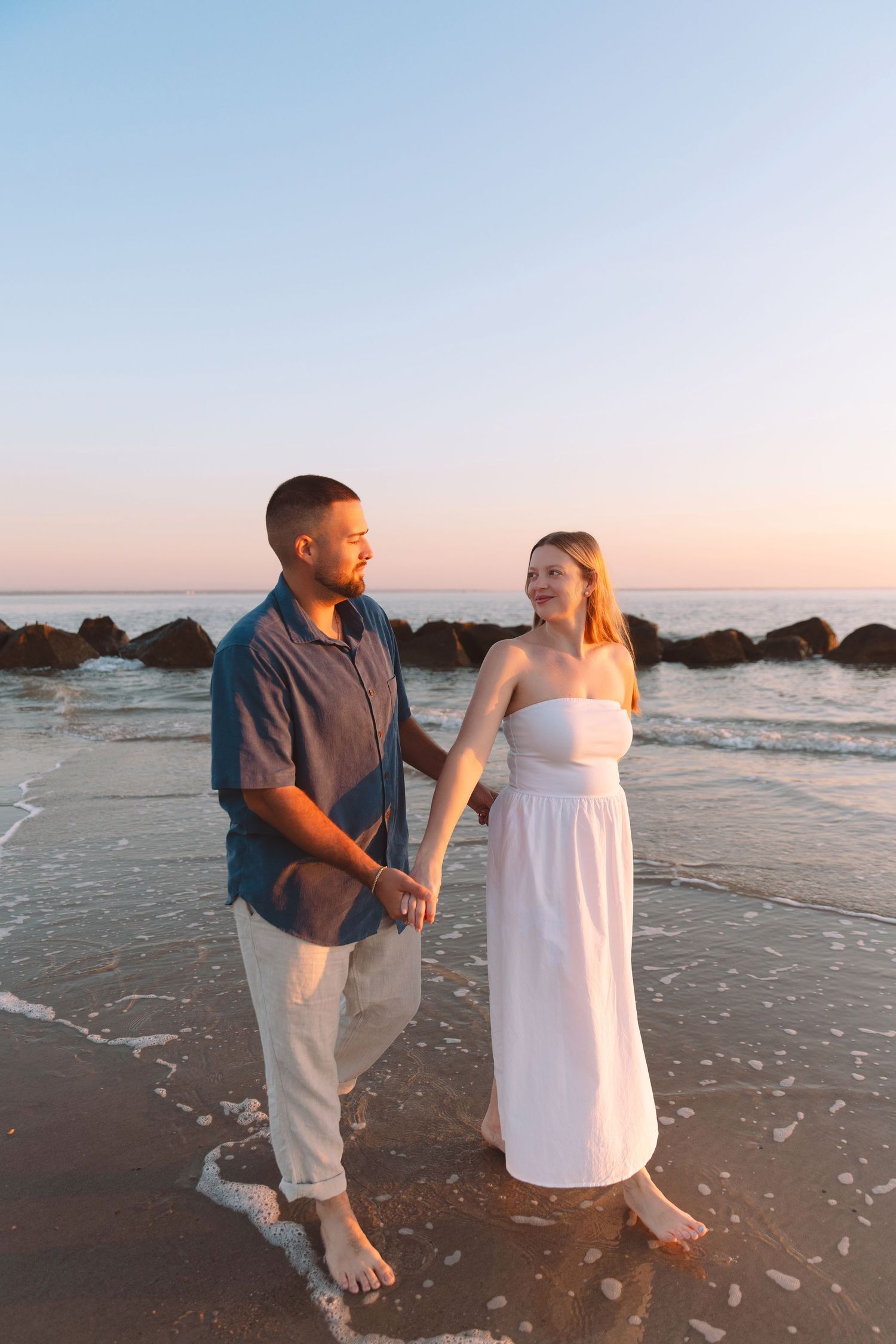 Couple holding hands walking barefoot on beach at sunset.