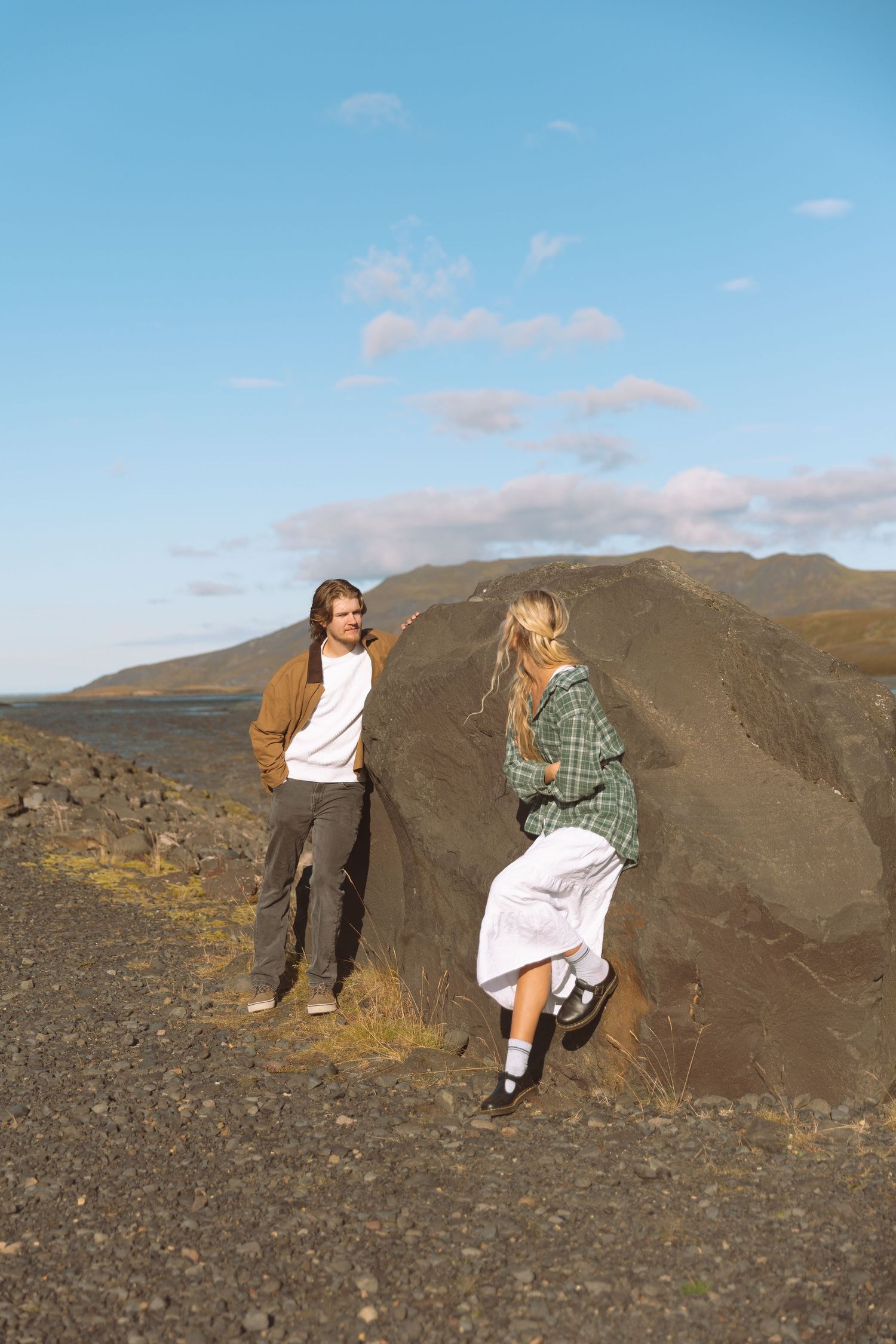 A young couple stands near a large volcanic rock on a barren highland landscape under a clear blue sky.