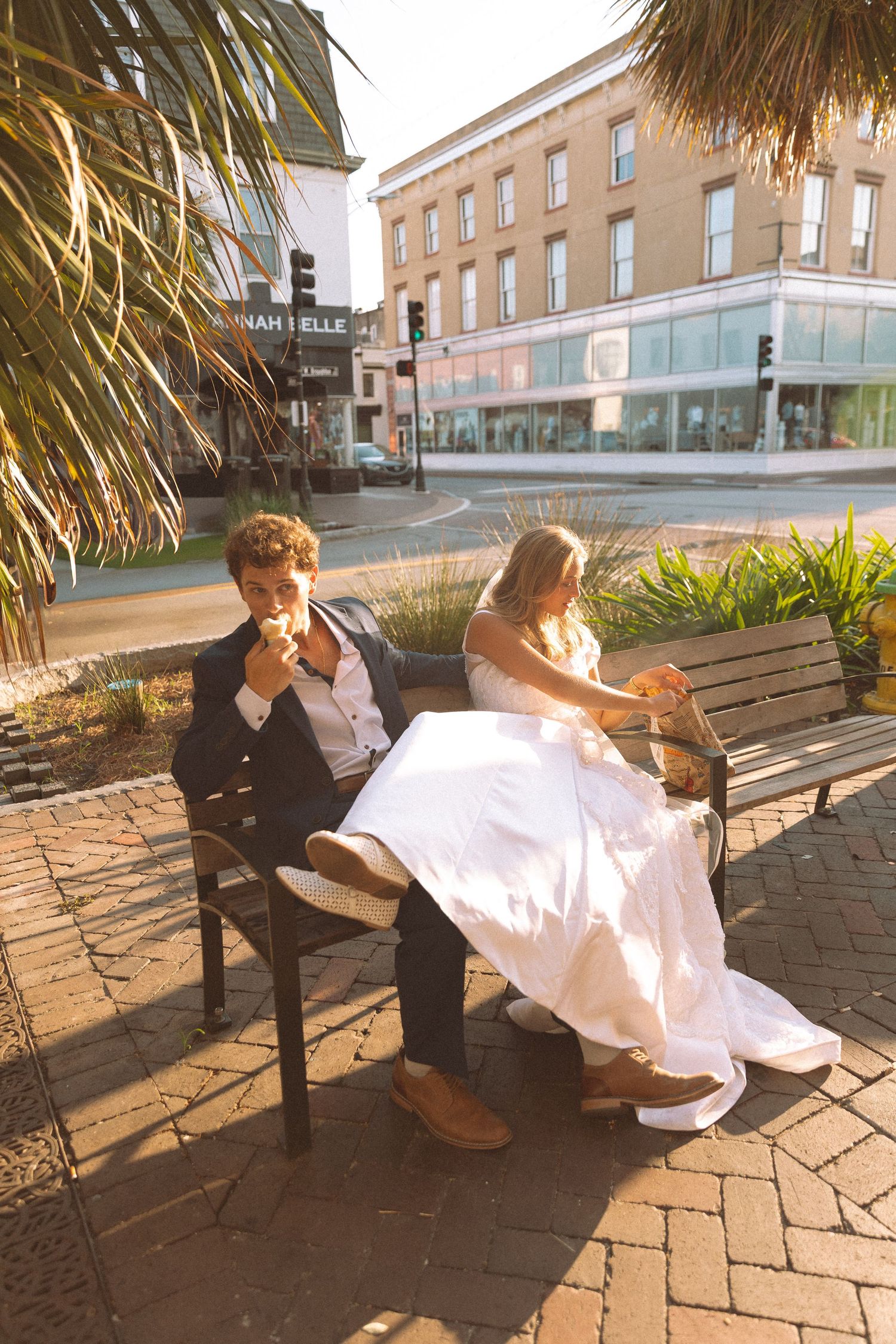 Bride and groom in wedding attire sit on bench eating outdoors in sunny urban setting.