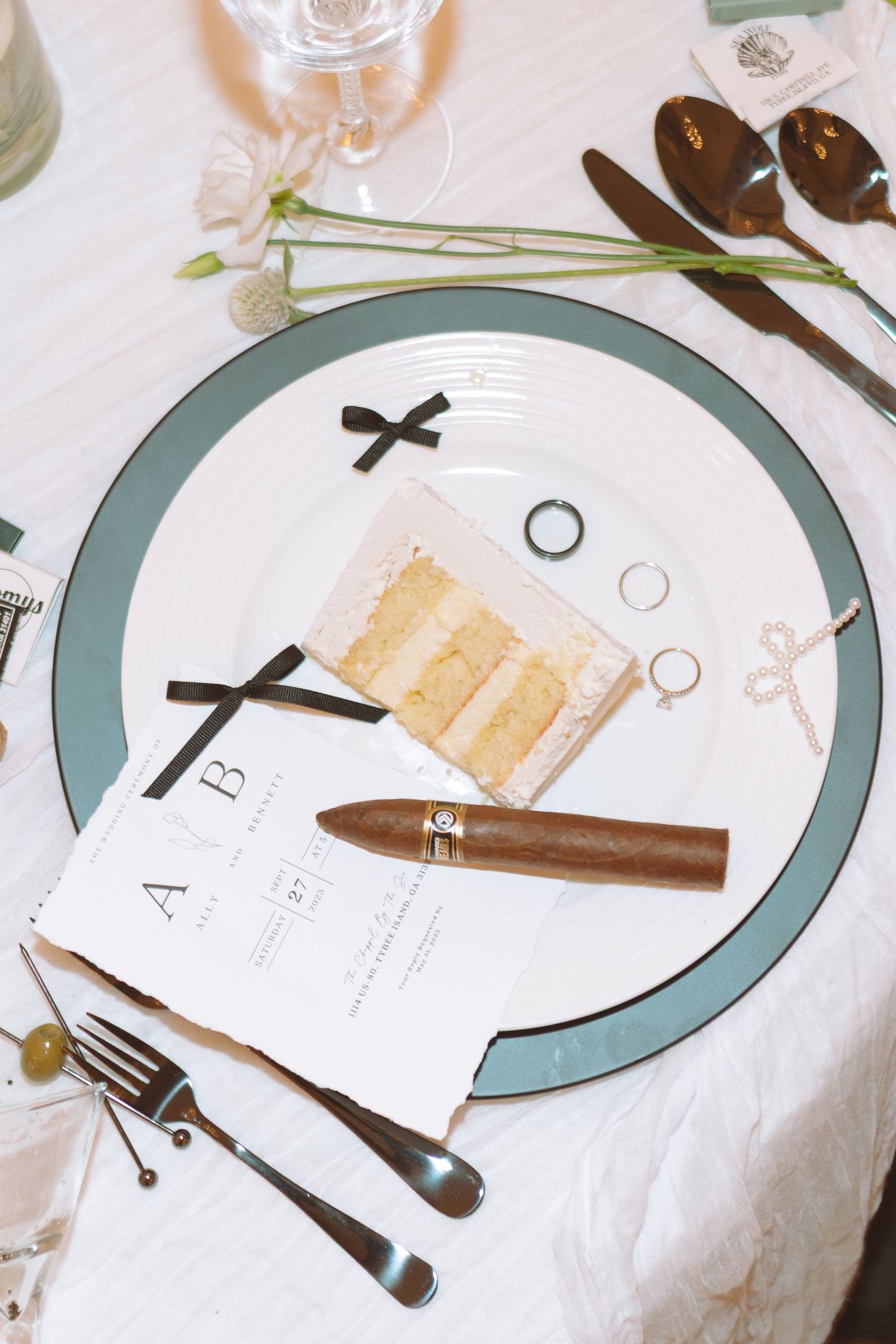 An elegant wedding place setting with a teal-rimmed plate, cigar, cake slice, rings, and monogrammed menu card.