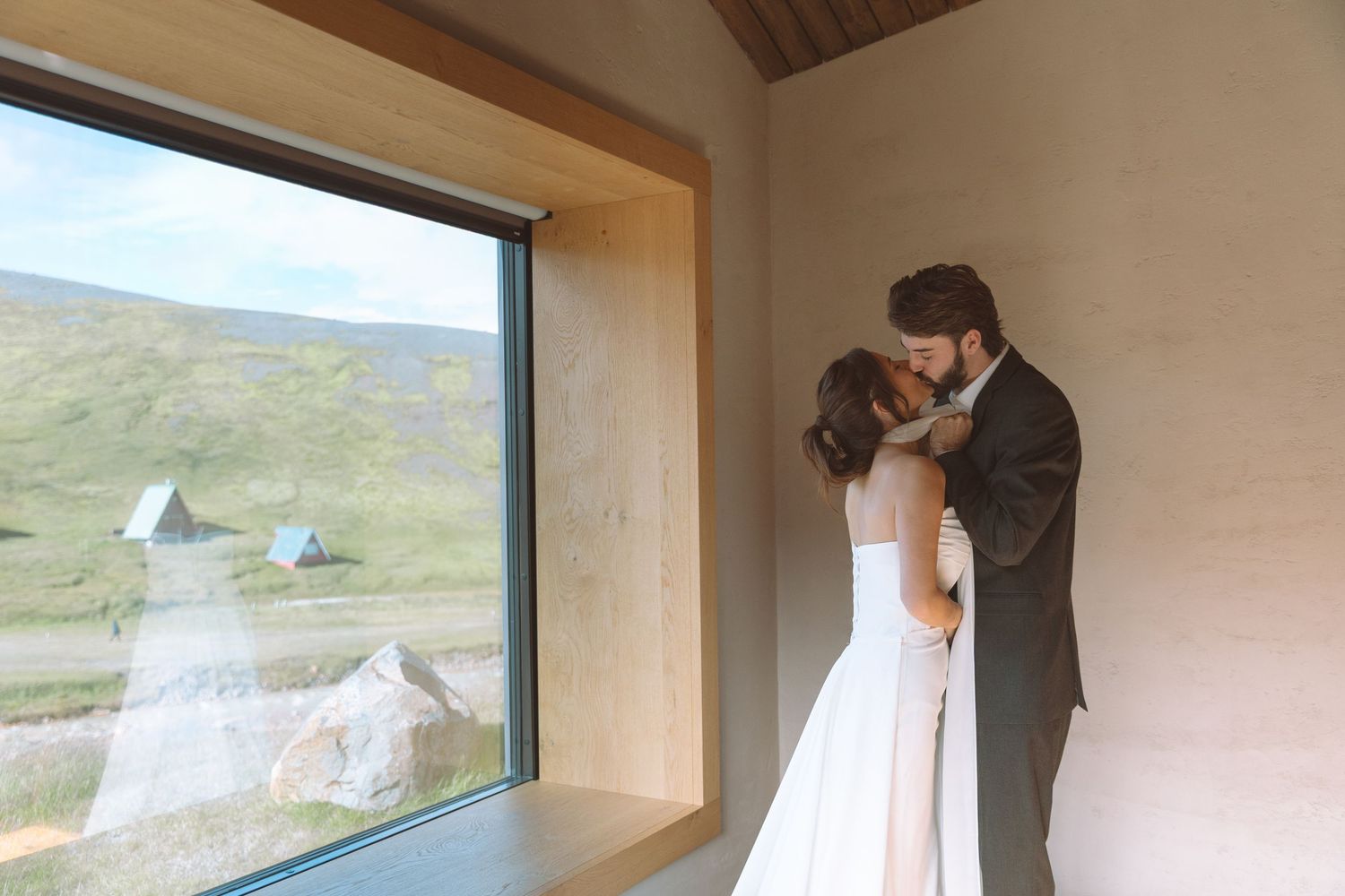 Wedding couple kissing by a large picture window with scenic green hillside views.