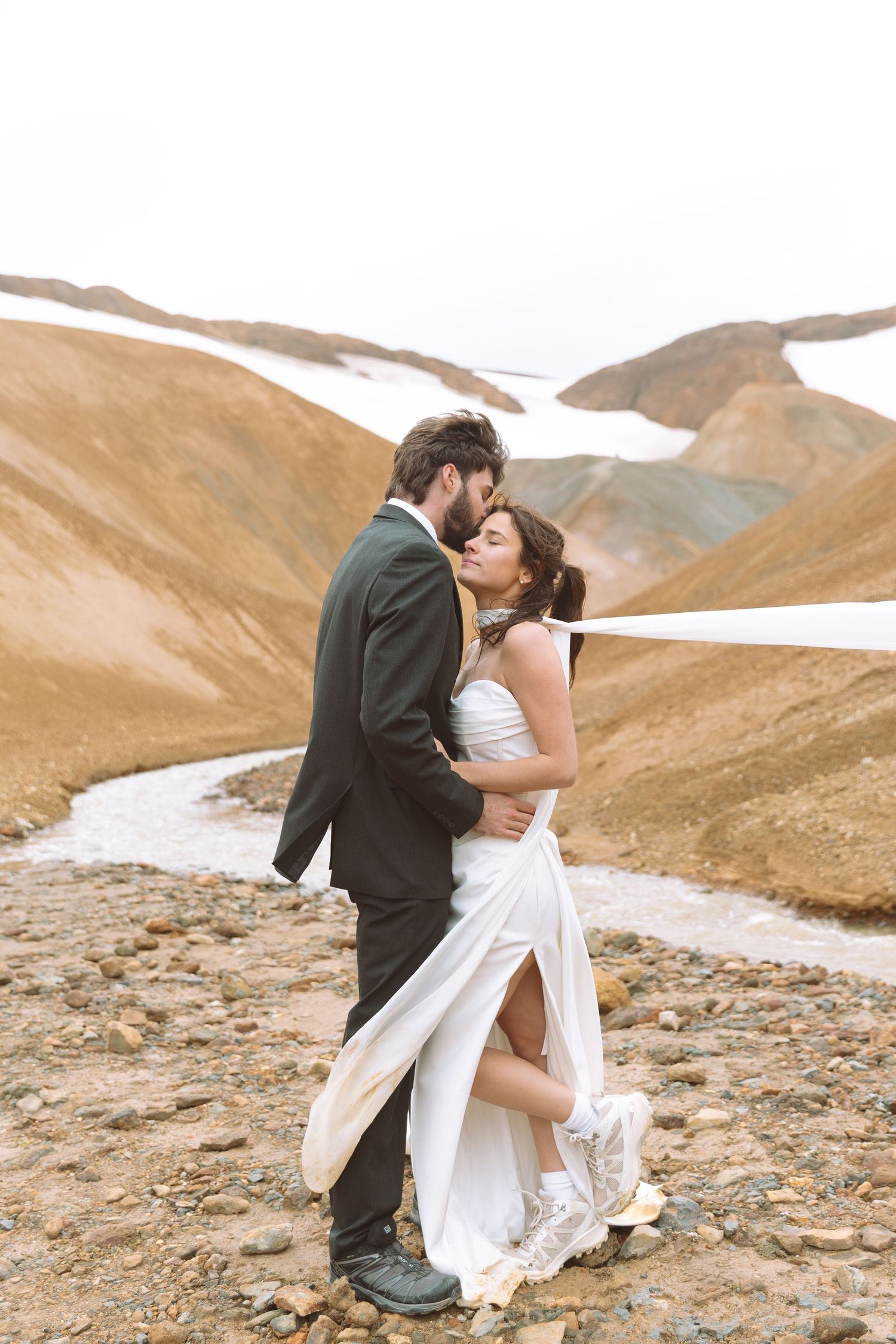Wedding couple shares a tender embrace on rocky ground with dramatic snow-streaked brown mountains behind them.