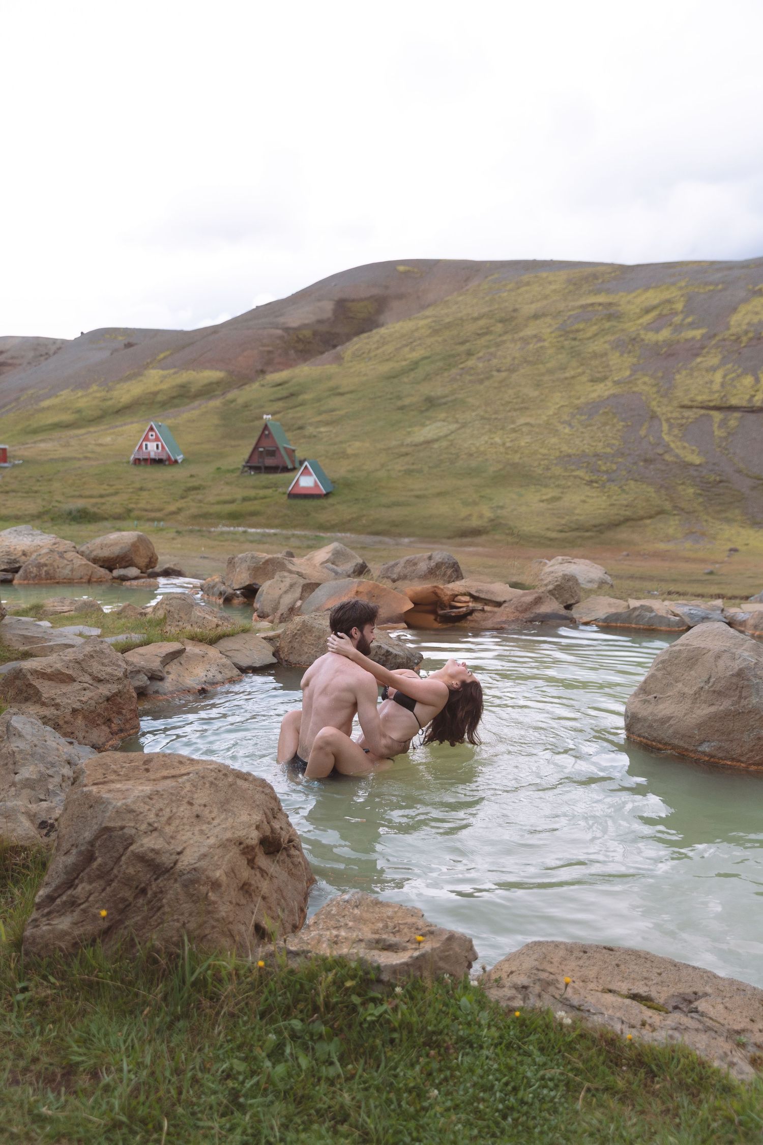 Romantic couple embracing in a natural hot spring with green Icelandic hills backdrop.