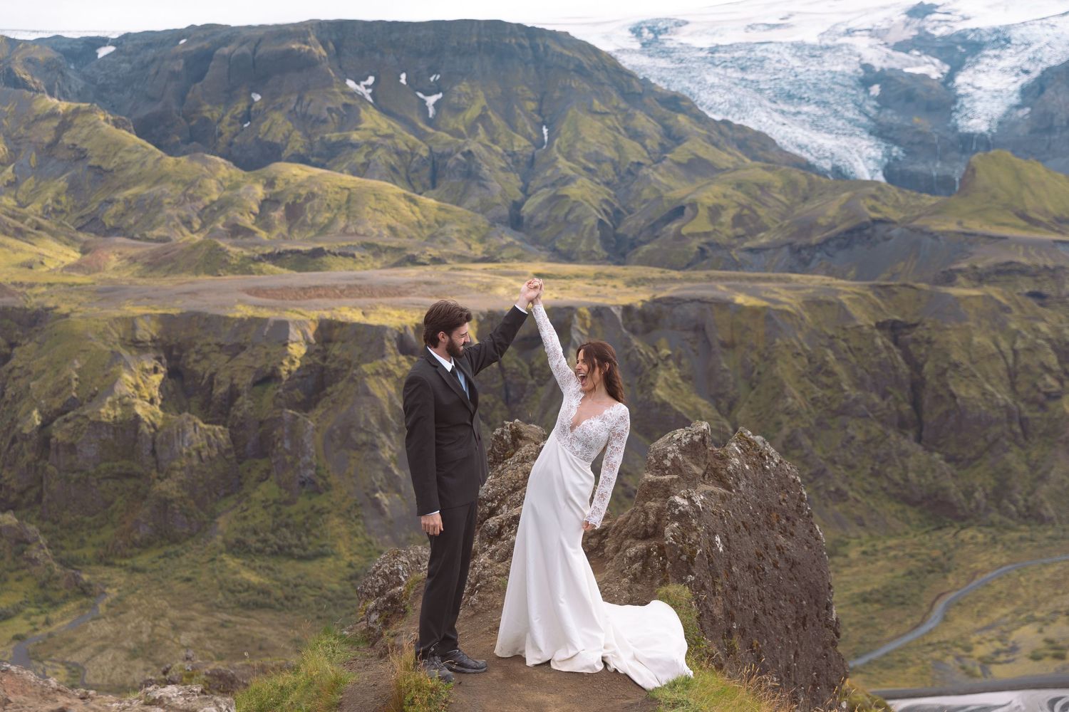 Joyful newlyweds celebrate on rocky Icelandic mountain peak with glacier backdrop after elopement ceremony.