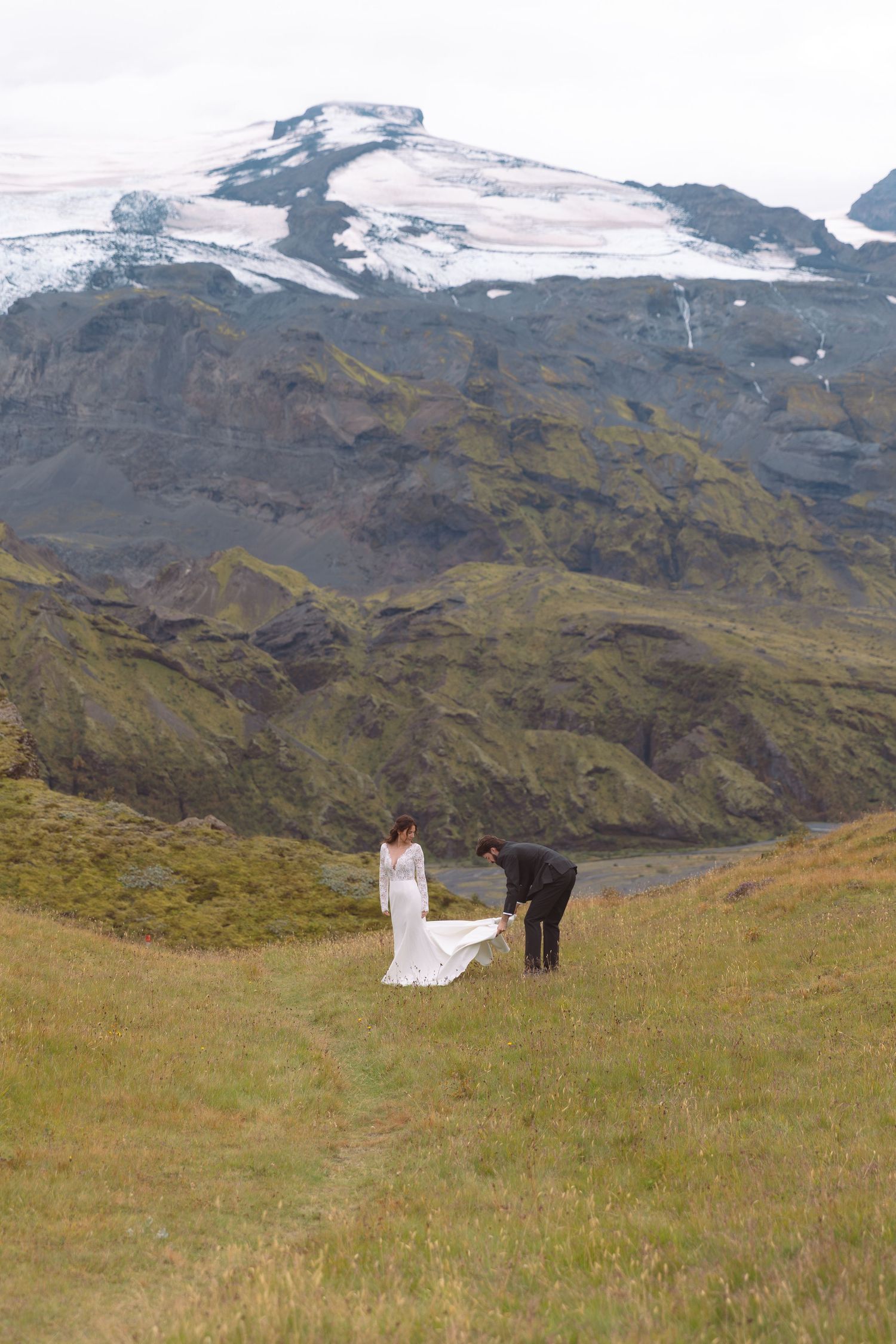 Groom arranging bride's wedding dress train on green hillside with massive snow-capped Icelandic volcano behind.